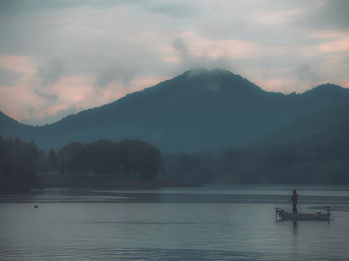 朝の湖畔でボートと釣り人、遠くに山並みが霞む静かな風景