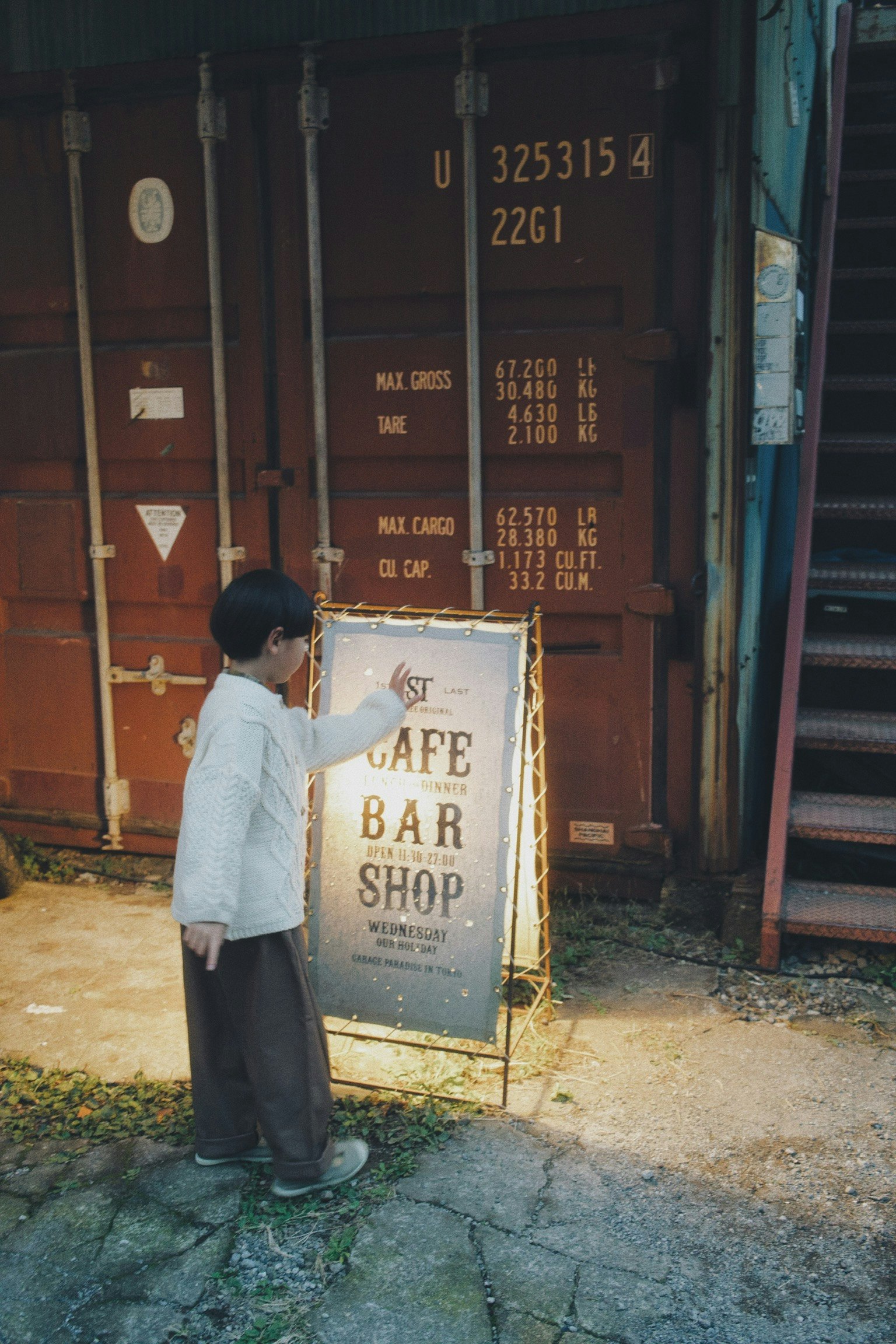 Child holding a cafe bar shop sign with shipping containers and stairs in the background