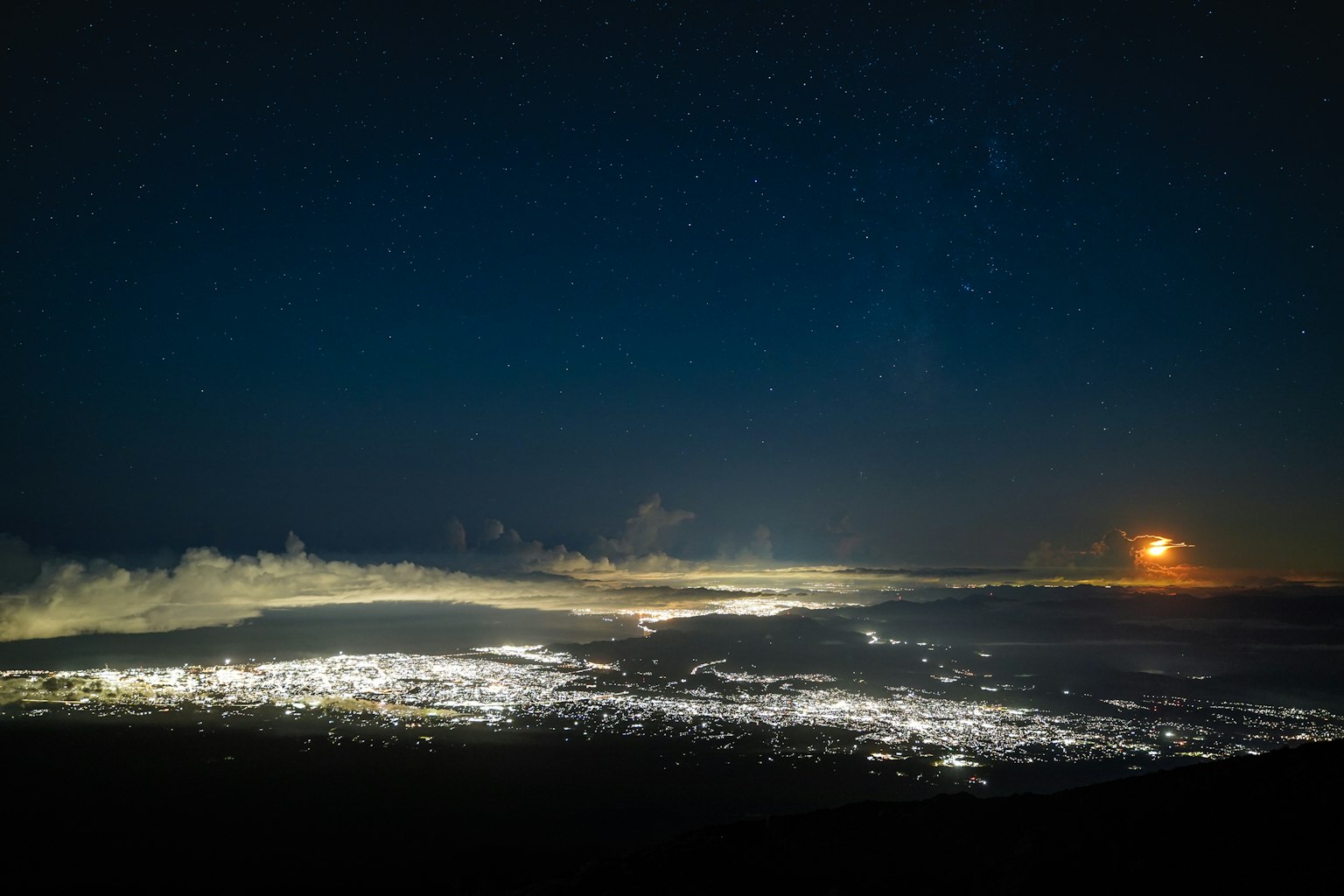 Pemandangan kota yang diterangi di malam hari di bawah langit berbintang dengan bulan yang jauh