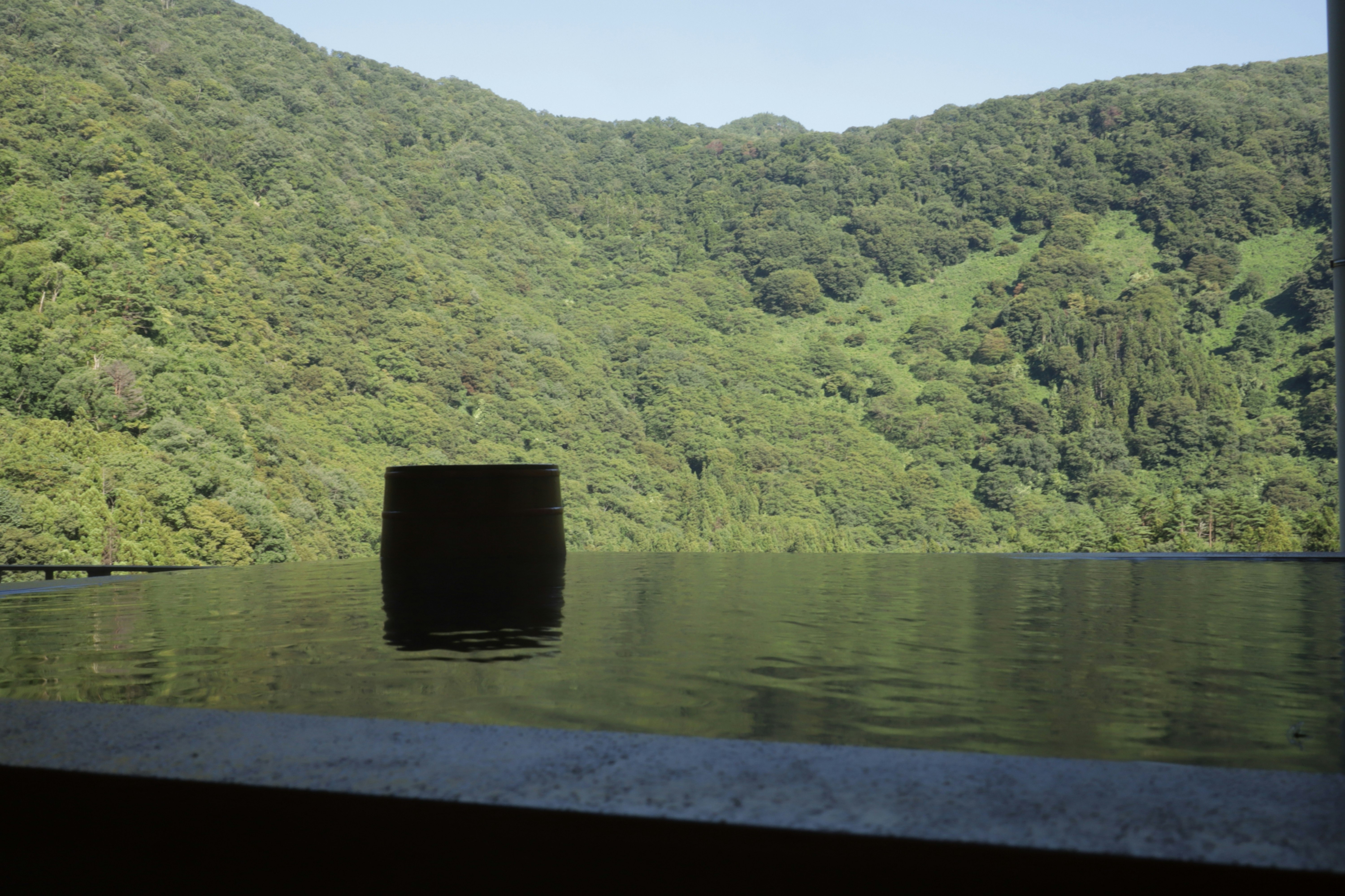 緑の山々に囲まれた湖面に浮かぶ黒い筒状オブジェのある風景