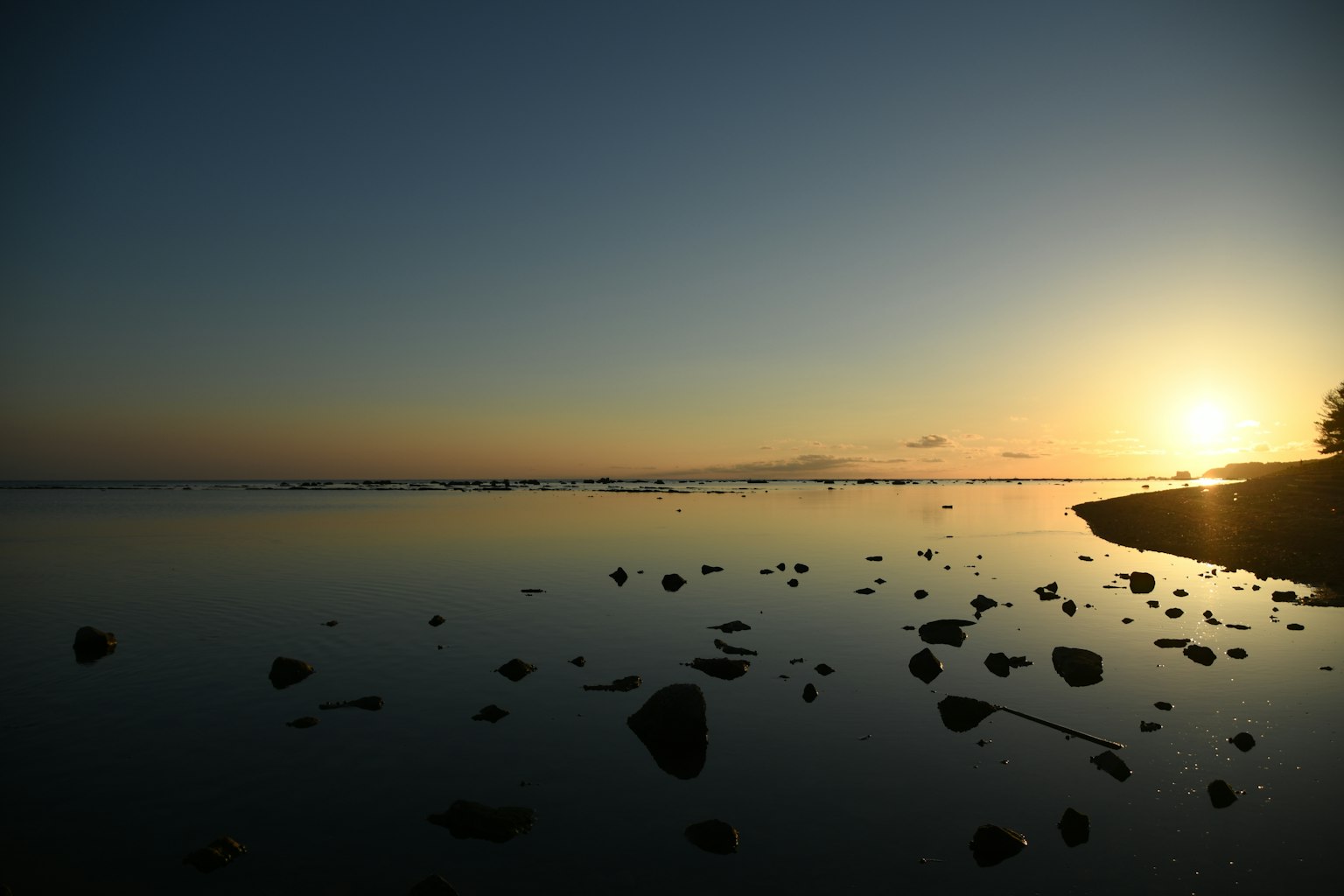 Pemandangan laut tenang dengan refleksi matahari terbenam dan tepi pantai berbatu