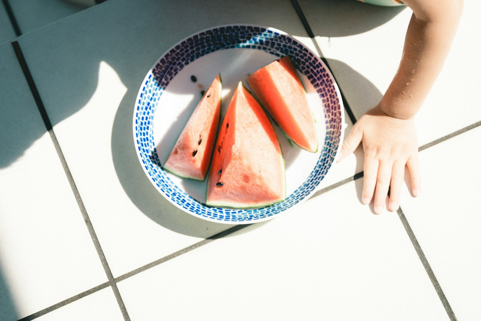Rebanadas de sandía en un plato sobre baldosas blancas con una mano de niño
