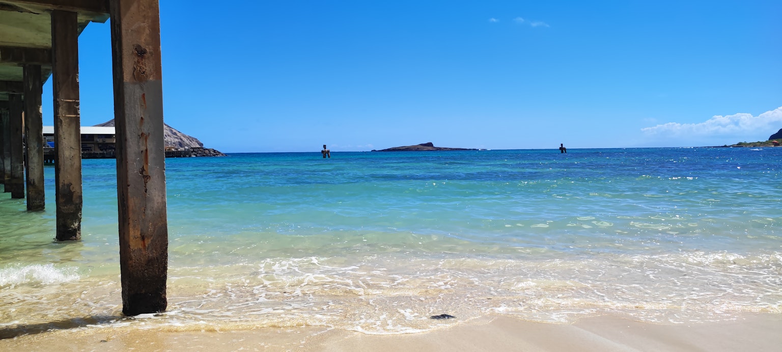 Scenic view of clear blue ocean and sandy beach with wooden pillars of a pier