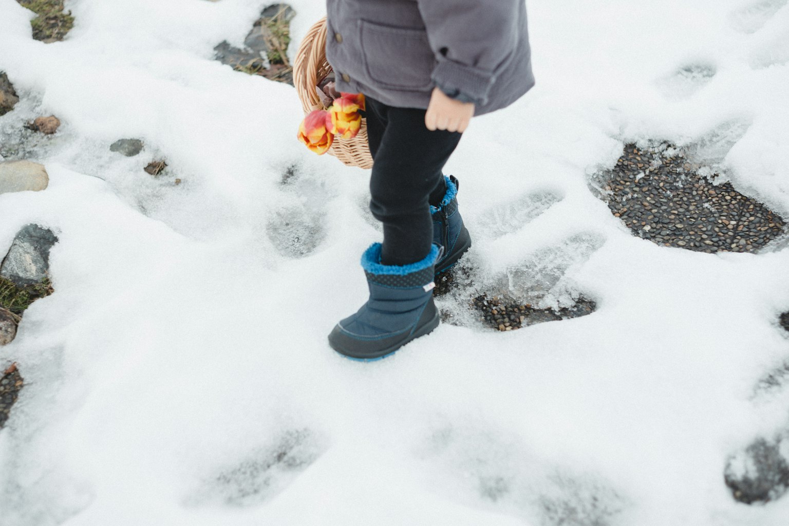 孩子穿着蓝色靴子在雪上行走，手里拿着五颜六色的花