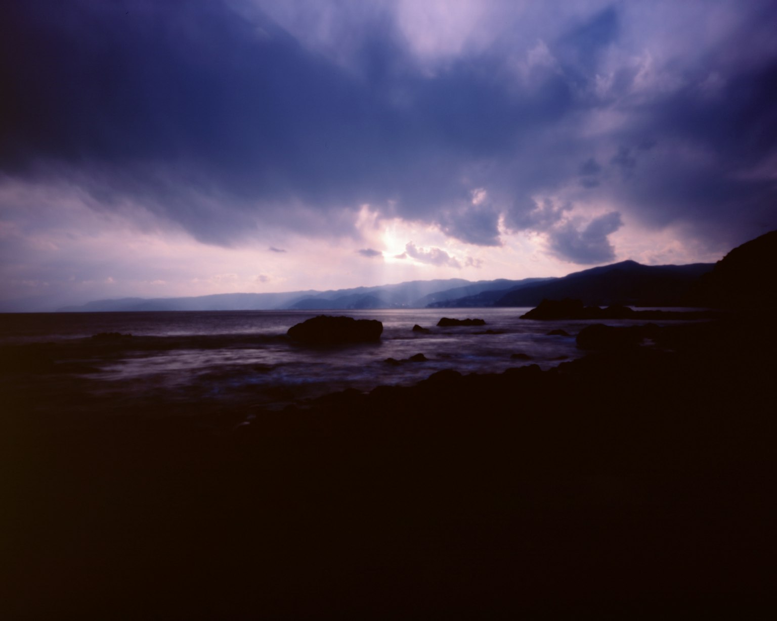 暗い海と岩がある風景で、曇り空からわずかな光が差し込む