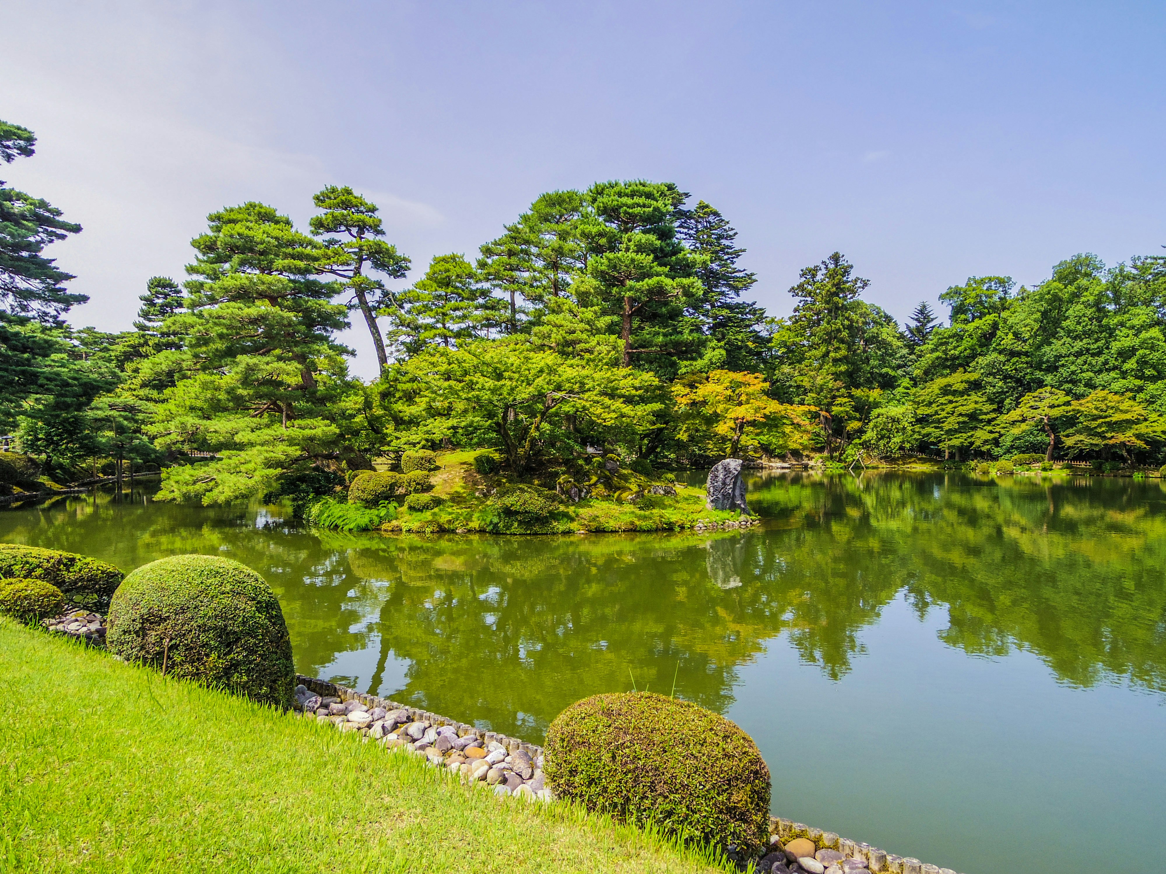 池のほとりに並ぶ刈り込まれた低木と苔むした石のある庭園の景色