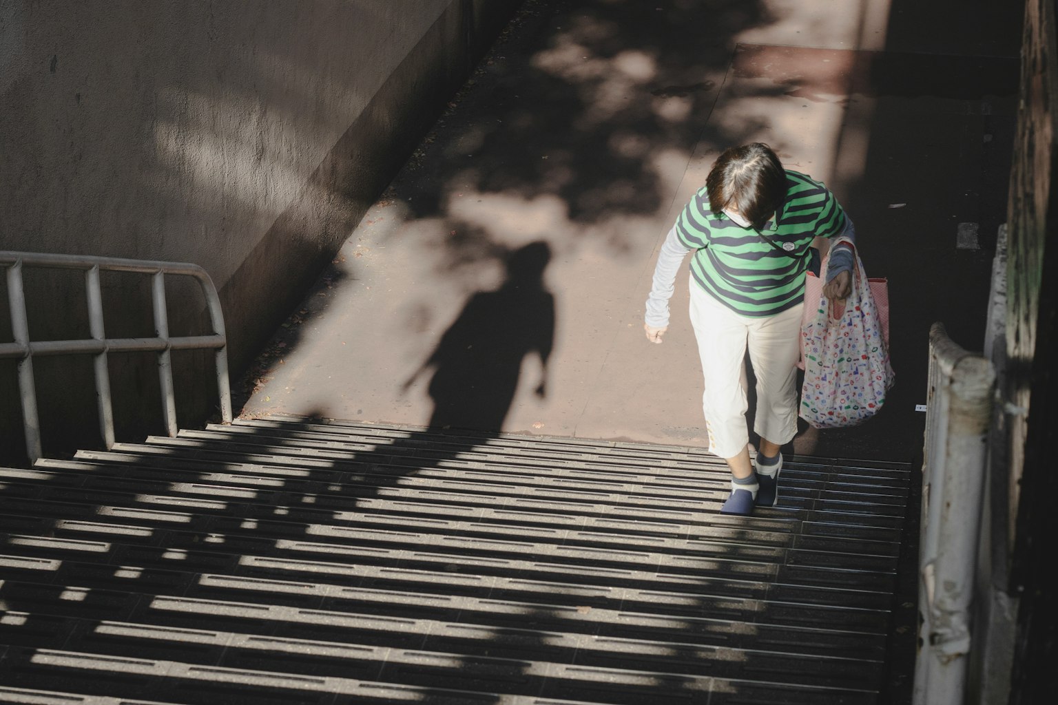Woman descending stairs wearing a green striped shirt and white pants with shadows cast by sunlight