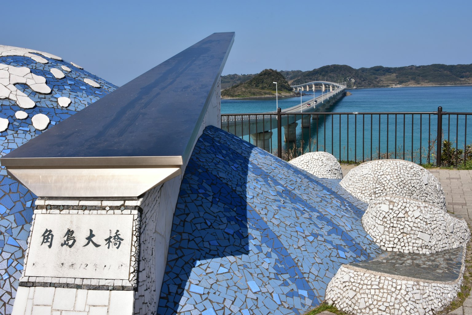 Küstenansicht mit blauer Mosaikskulptur und Brücke im Hintergrund