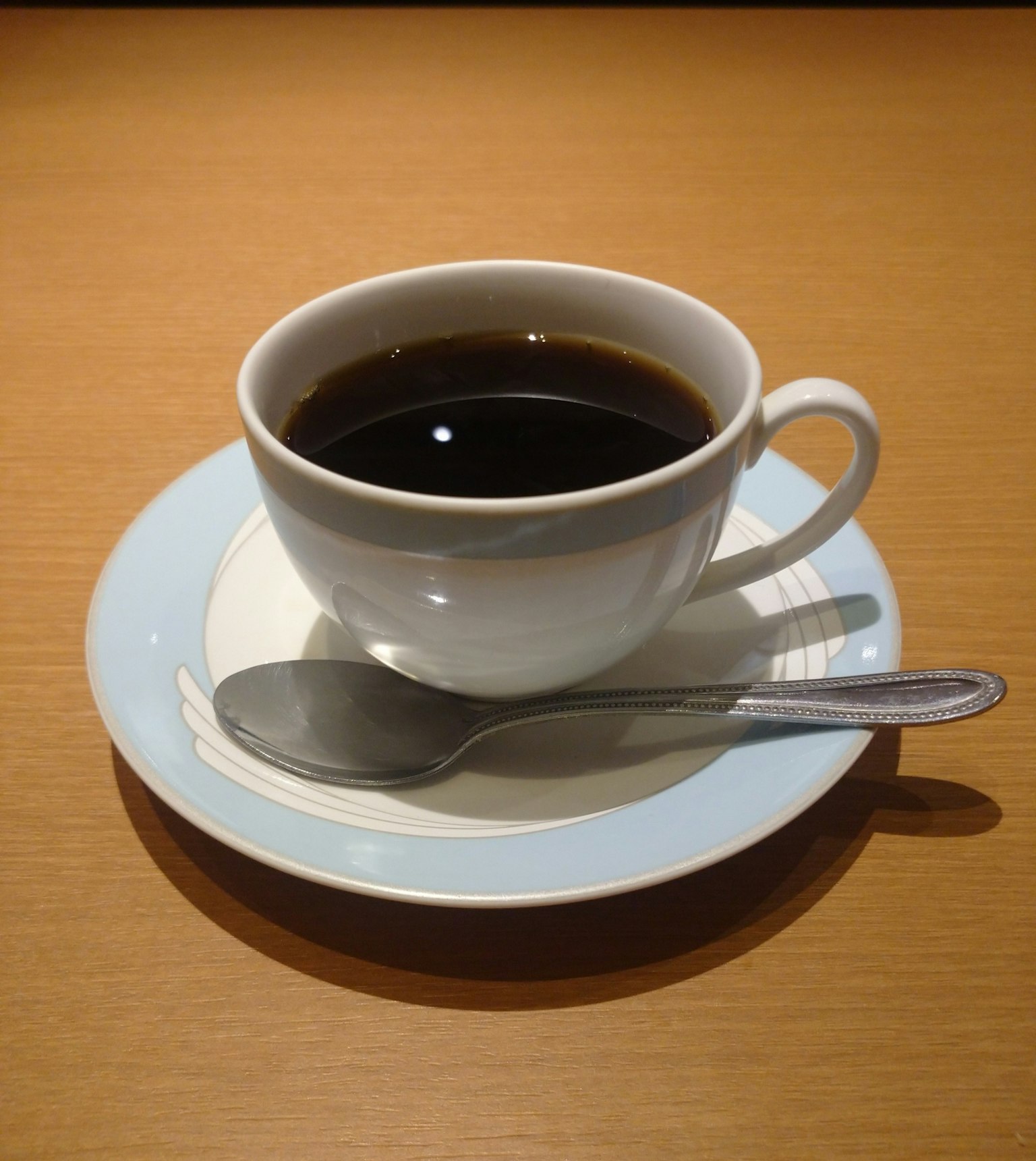 Kopi hitam dalam cangkir putih dengan sendok perak di atas piring berpinggiran biru muda