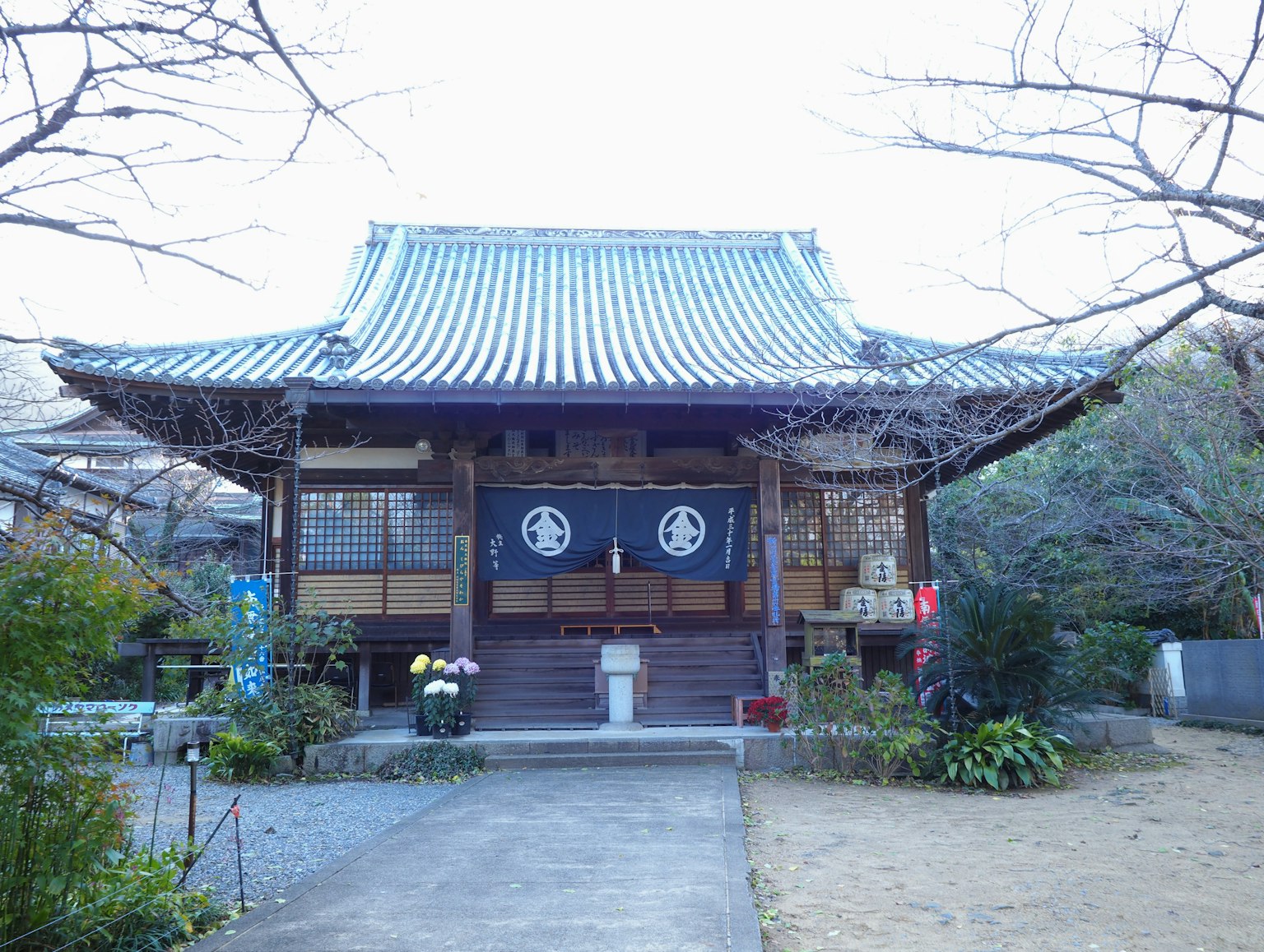 Exterior de un templo japonés tradicional con techo de tejas azules y puerta blanca rodeada de vegetación