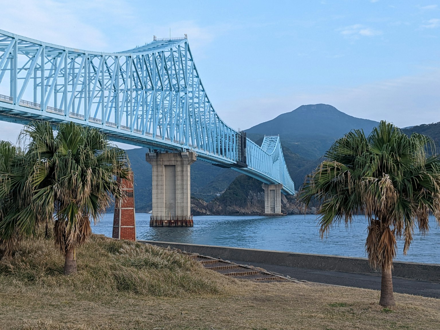 A blue bridge spans over a blue sea with palm trees in the foreground
