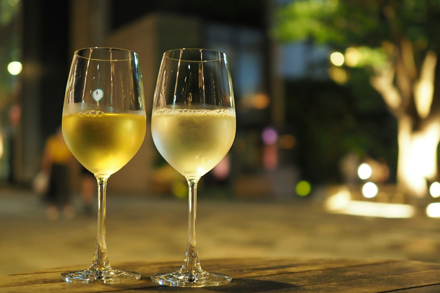 Due bicchieri di vino riempiti di vino bianco con luci sfocate e un albero sullo sfondo