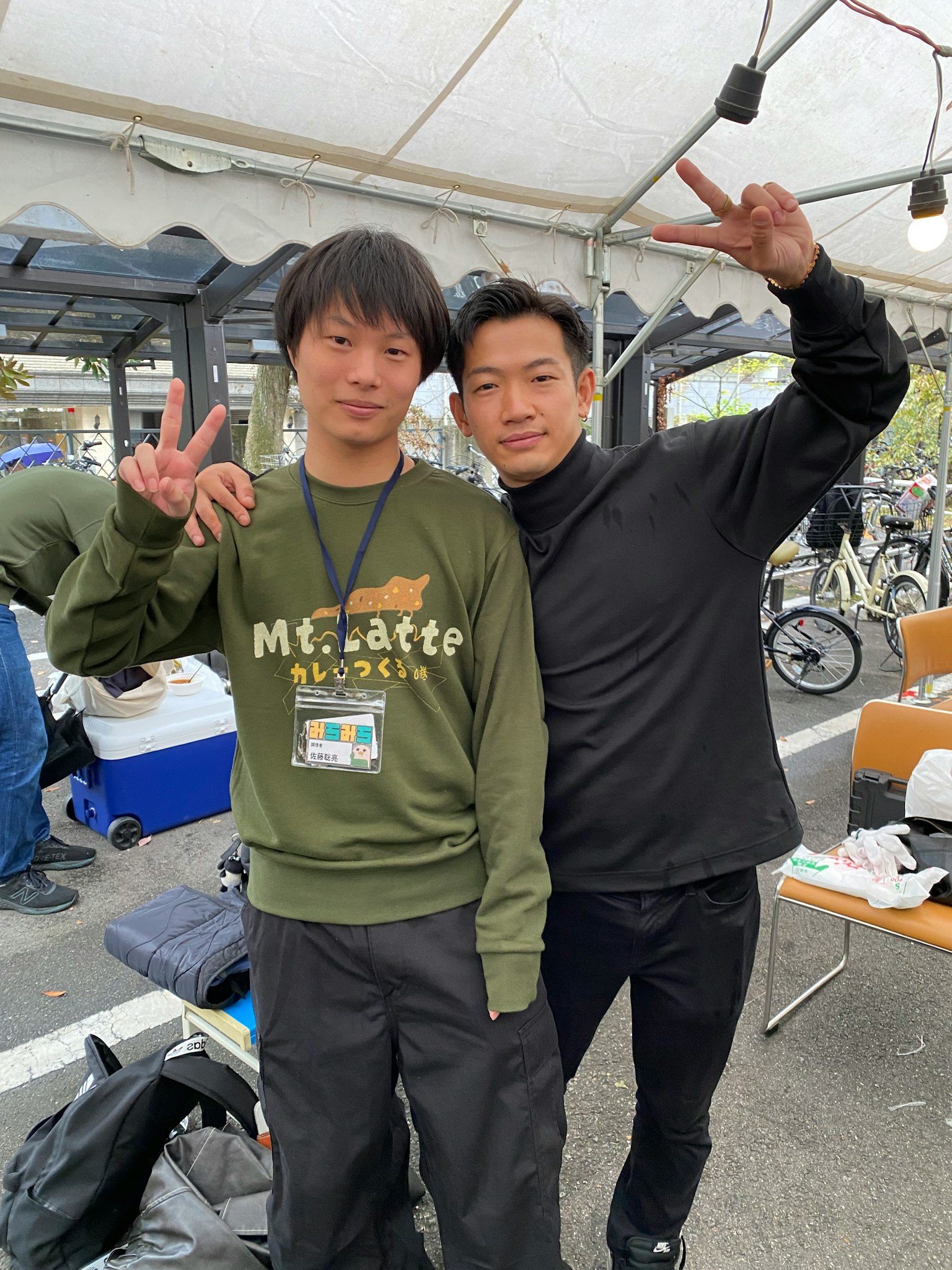 两名男子摆出和平手势一名穿着绿色卫衣另一名穿着黑色高领衫背景中有帐篷和椅子