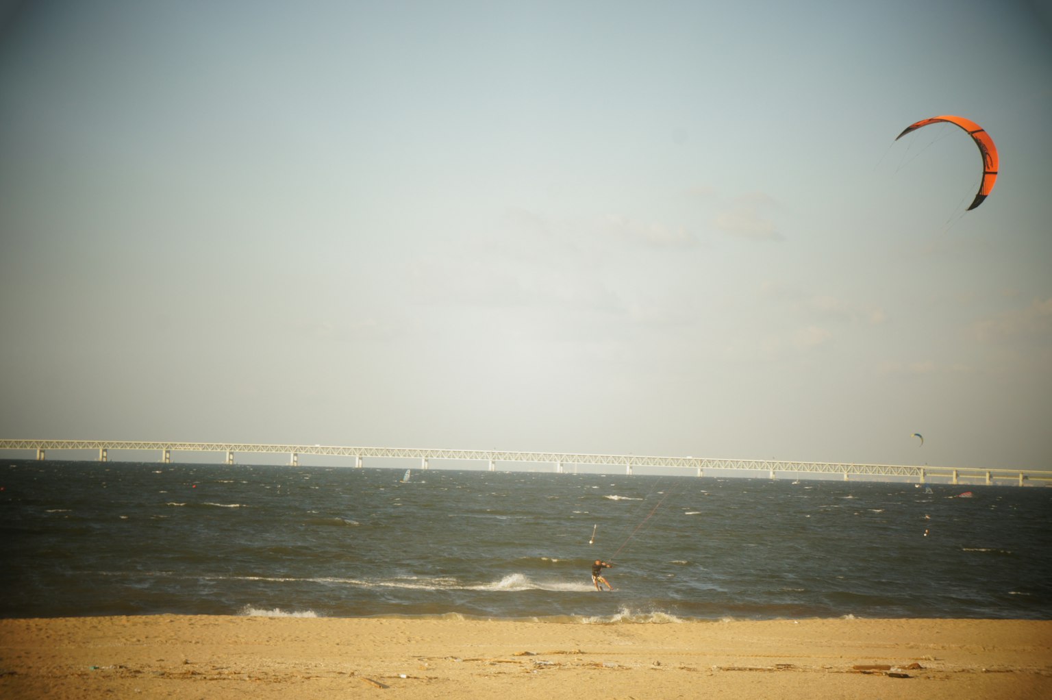 Eine Person beim Kitesurfen am Strand mit einer Brücke in der Ferne
