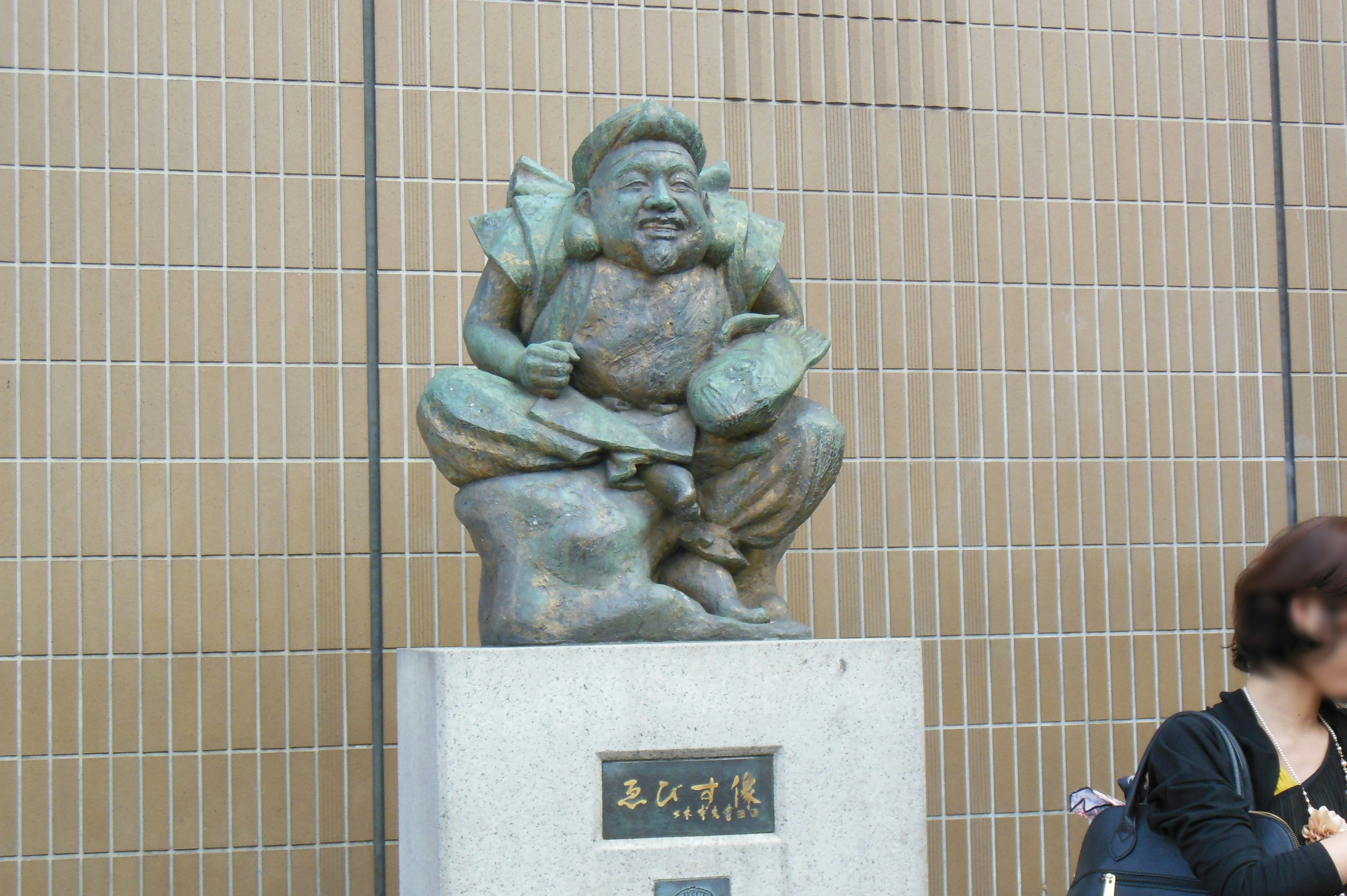 statue de lutteur de sumo en pierre devant un mur carrelé avec un passant