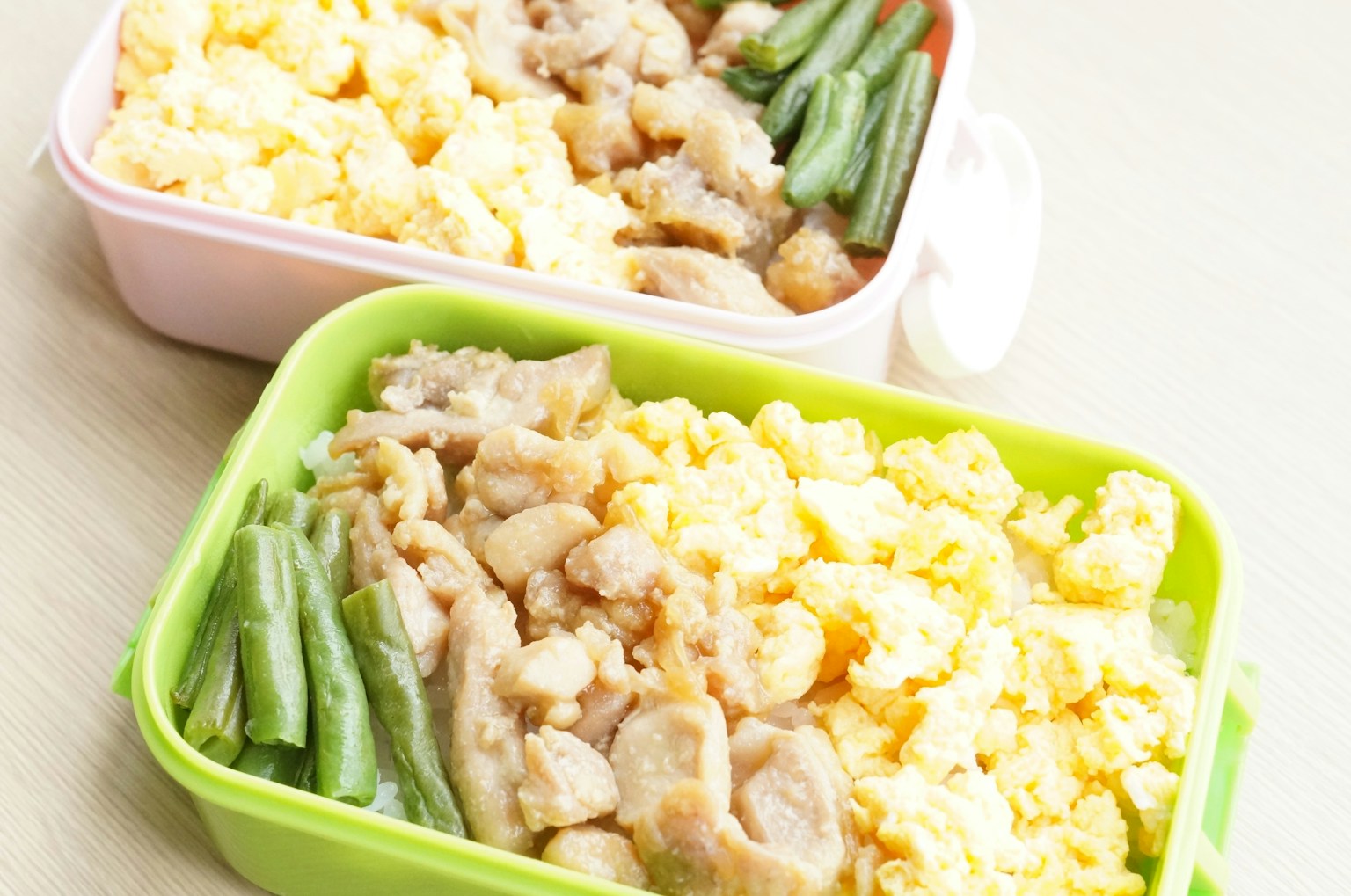 Kotak bento hijau dan merah muda diisi dengan telur orak-arik, ayam, dan kacang hijau