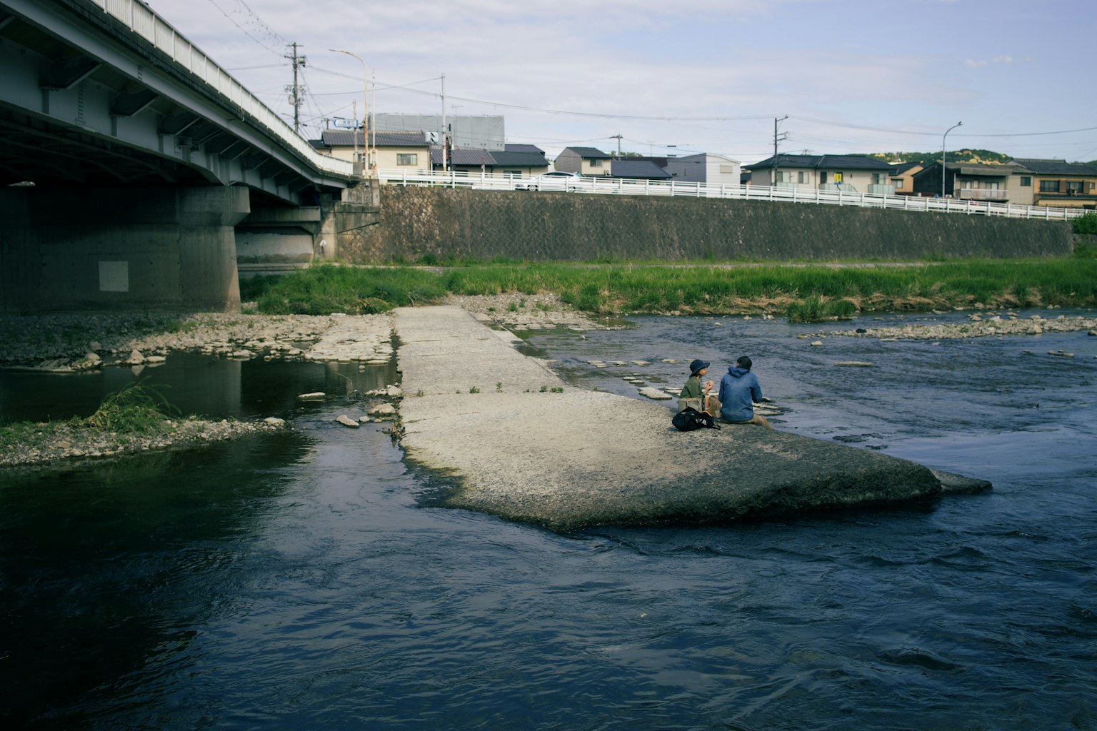 Two people sitting by the river on a small island under a bridge