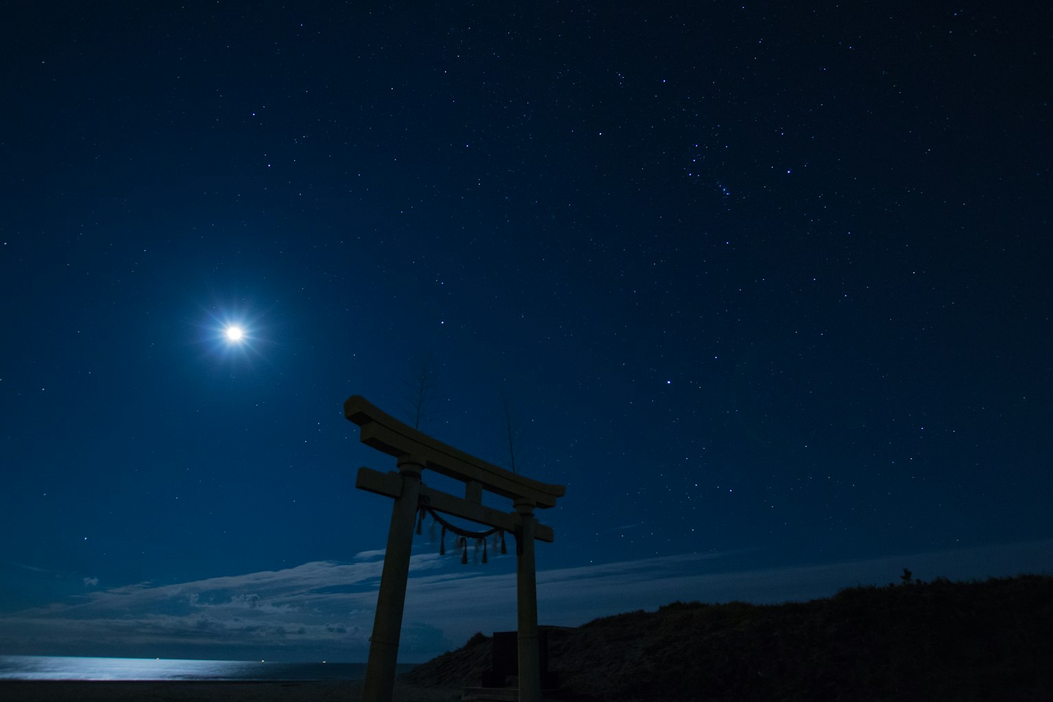 เสาโทริอิในเงายามค่ำคืนที่เต็มไปด้วยดาวพร้อมกับดวงจันทร์ที่สว่าง