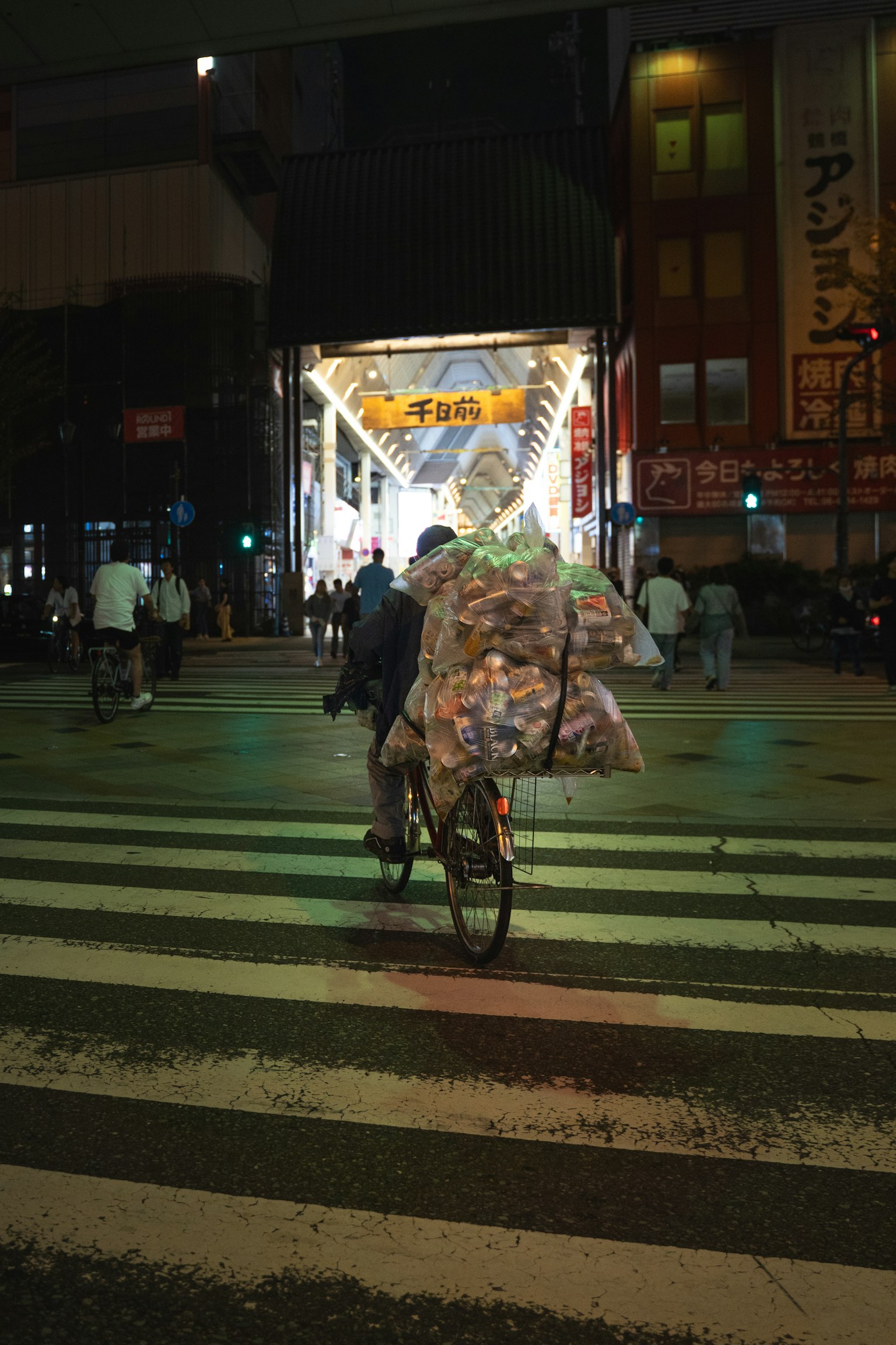 Un repartidor en bicicleta cruzando una intersección nocturna con bolsas apiladas en su bicicleta