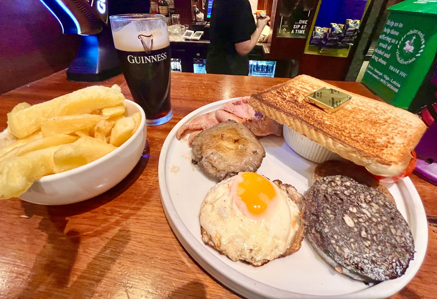 A plate with breakfast items including a sandwich fried egg sausage black pudding and a bowl of fries alongside a glass of Guinness