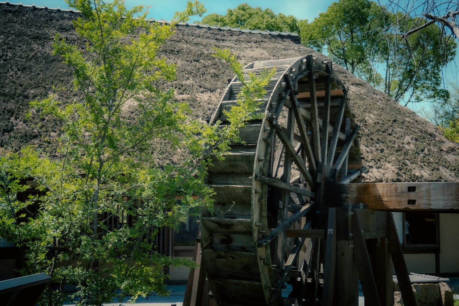 A traditional water wheel beside a thatched roof building surrounded by greenery