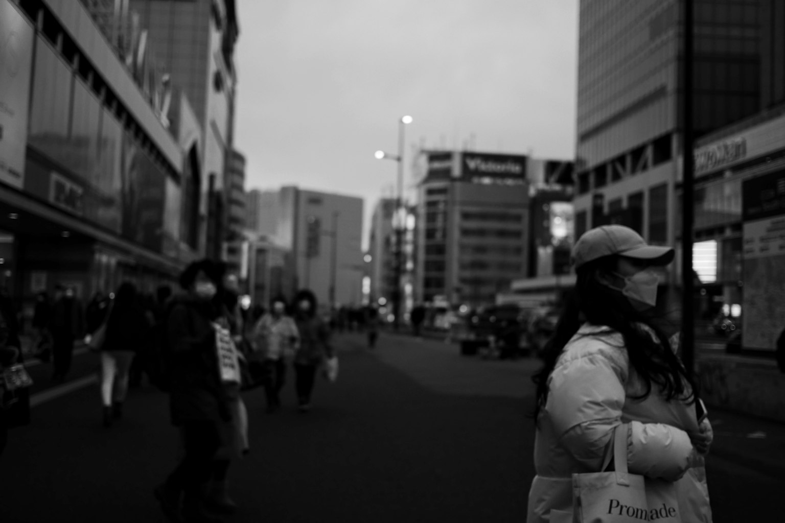 Una donna con una maschera si trova in una strada di città in bianco e nero con persone che camminano intorno