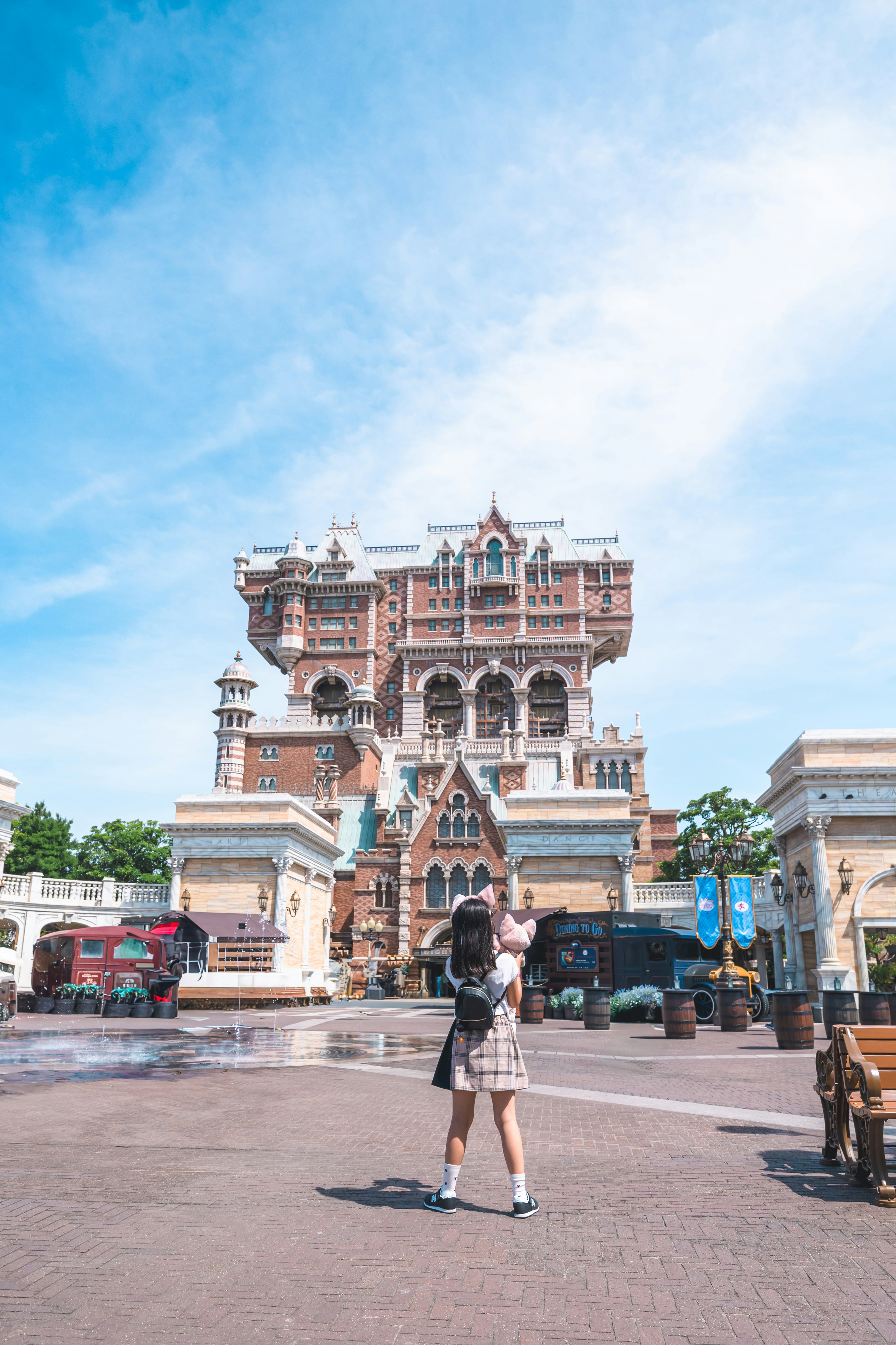 A woman stands in front of a towering structure in a theme park under a bright blue sky
