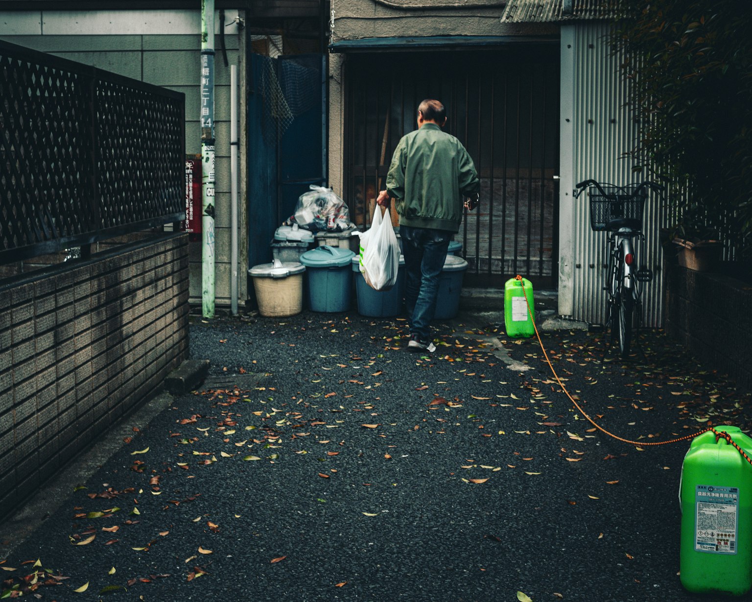 Un homme portant un sac poubelle dans une ruelle étroite avec un vélo et des conteneurs verts visibles
