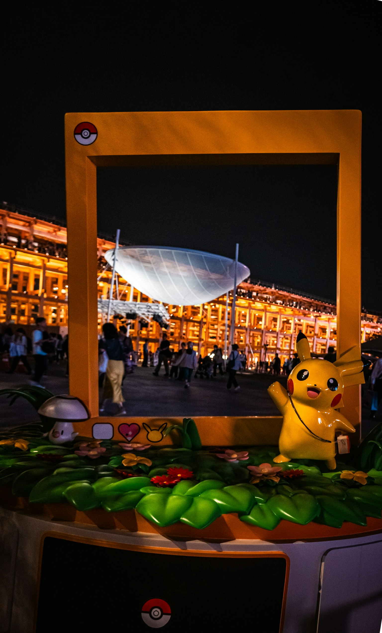 夜のイベントでピカチュウのフィギュアとポケモンのフレームがある背景に人々がいる