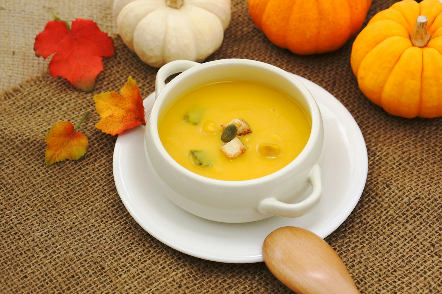 Soupe de potiron crémeuse servie dans un bol blanc entouré de citrouilles orange et de feuilles d'automne