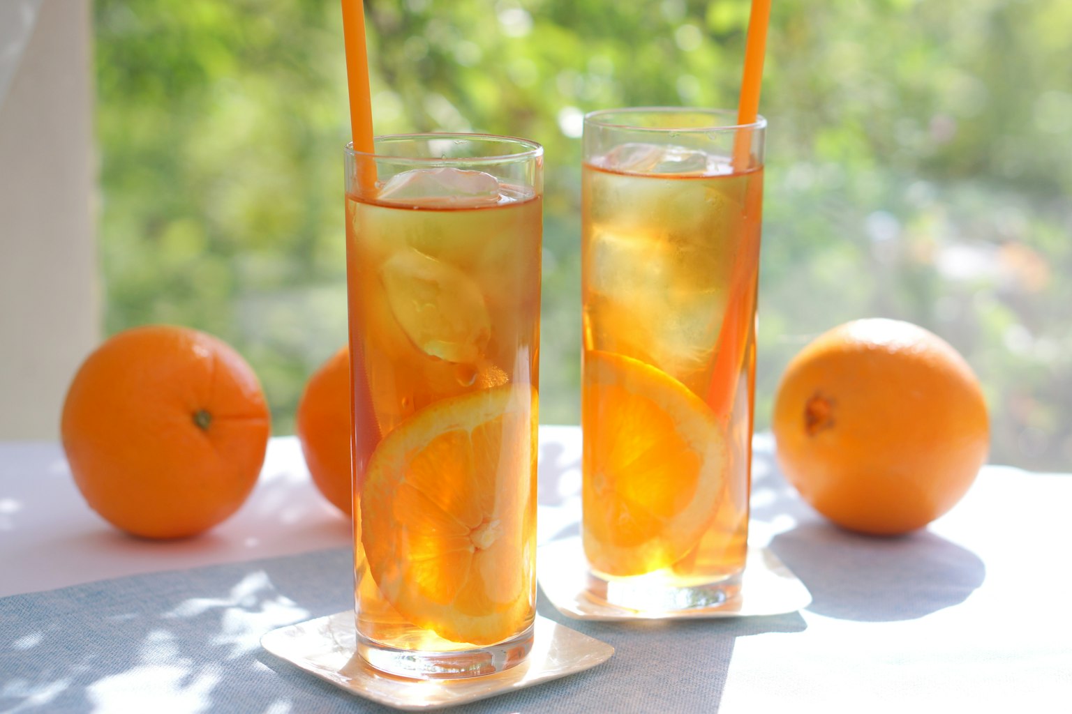 Two glasses of iced tea with orange slices and ice cubes in a bright setting with oranges nearby