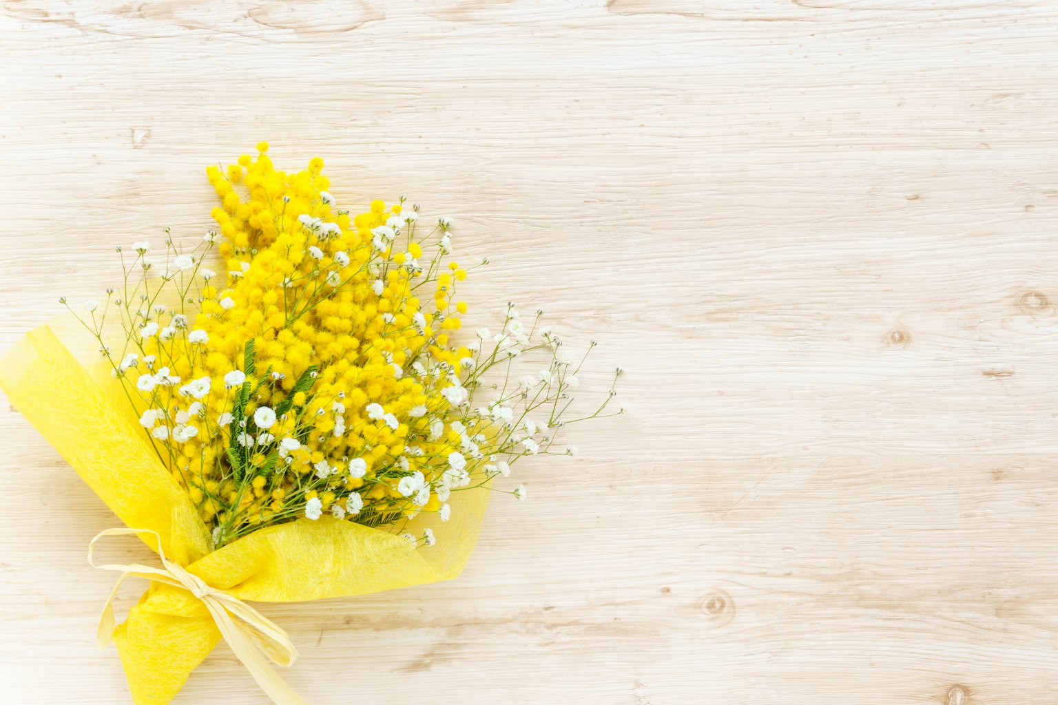 Một bó hoa mimosa vàng và hoa trắng được gói trong giấy vàng trên bàn gỗ