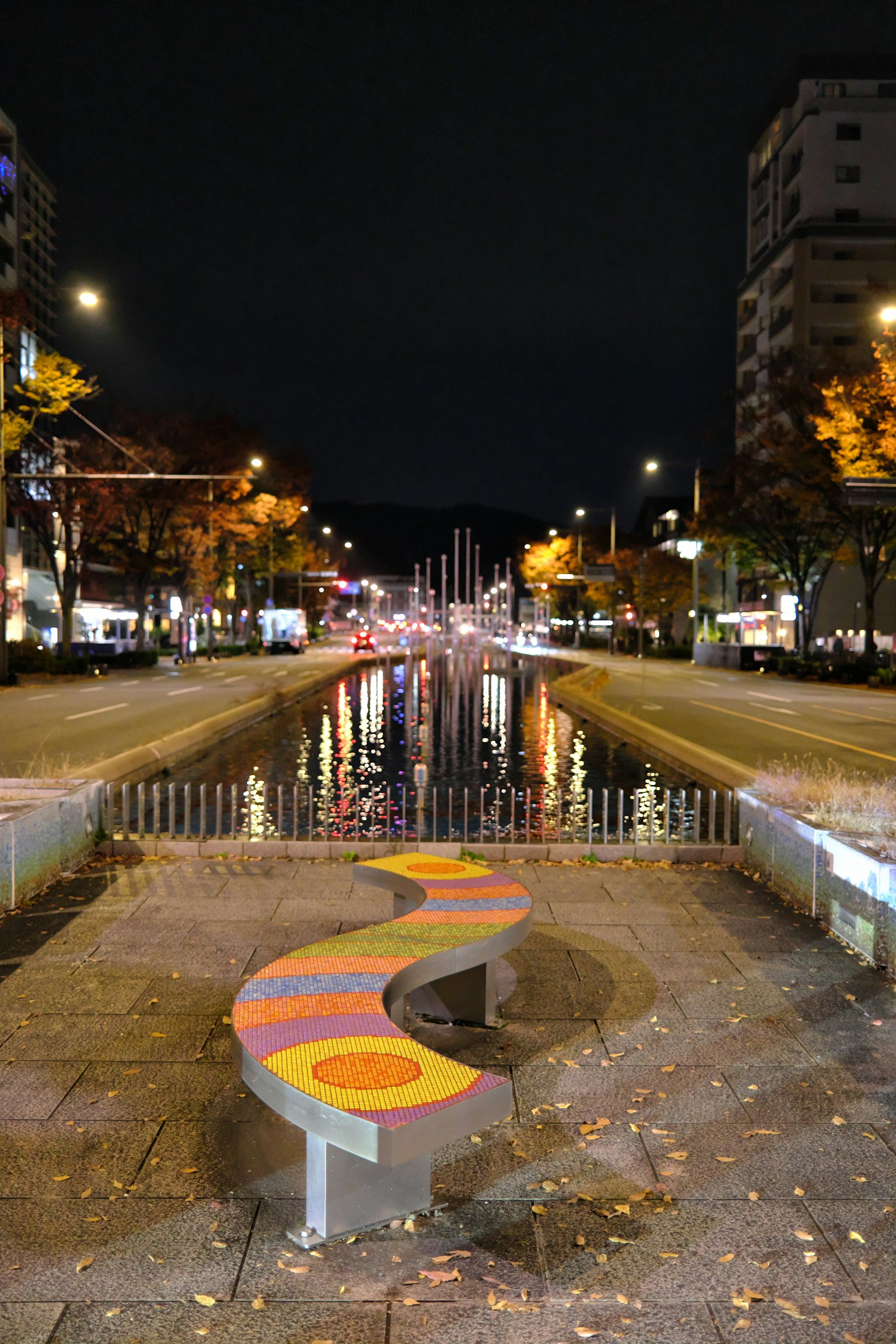 Una scena notturna con una panchina colorata accanto a un canale illuminato e una strada cittadina