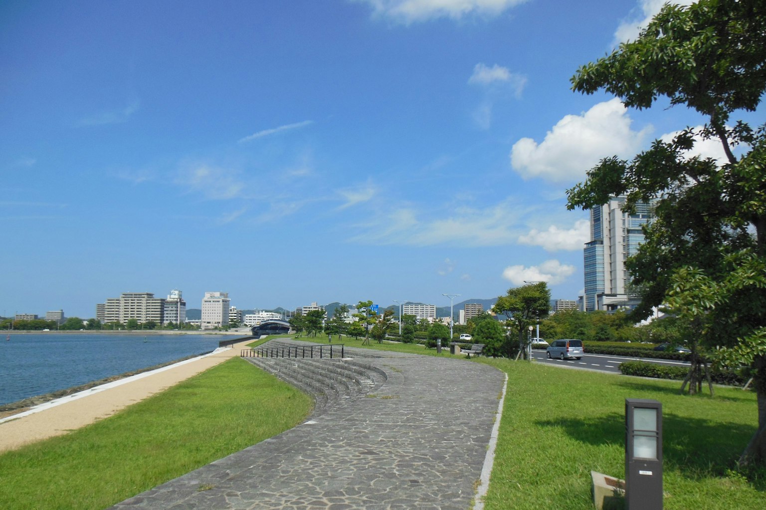 Scenic view of a riverside walkway under a blue sky featuring skyscrapers and lush greenery