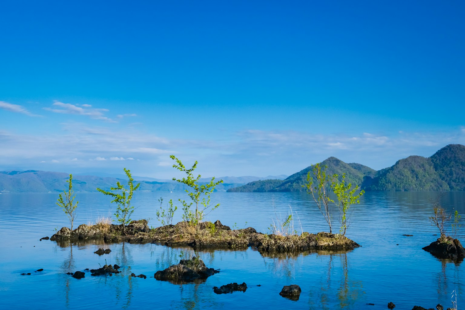 Pulau kecil dengan pepohonan dikelilingi air tenang dan pegunungan di bawah langit biru