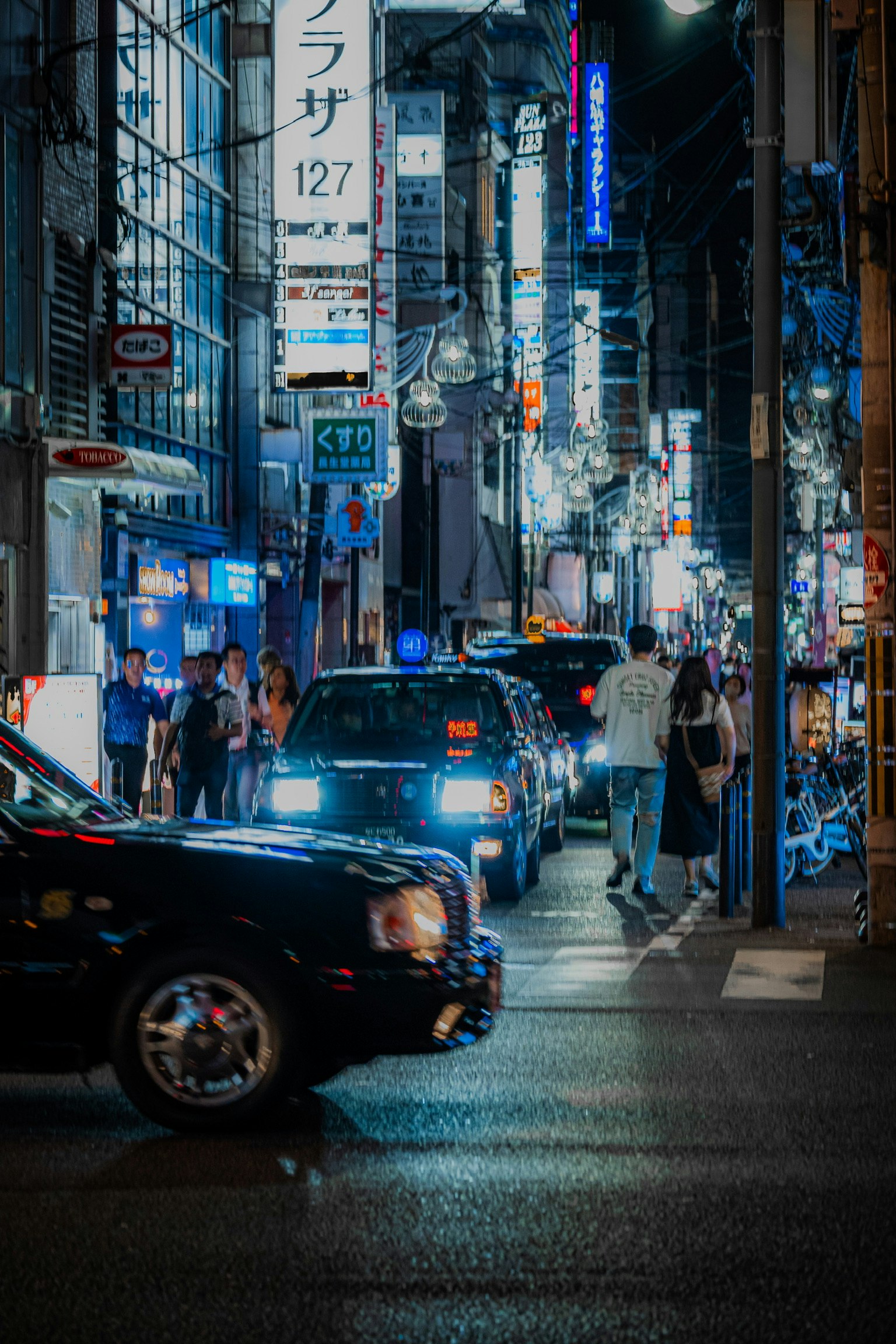 nächtliche Straßenszene mit Neon-Schildern, Fußgängern und Autos