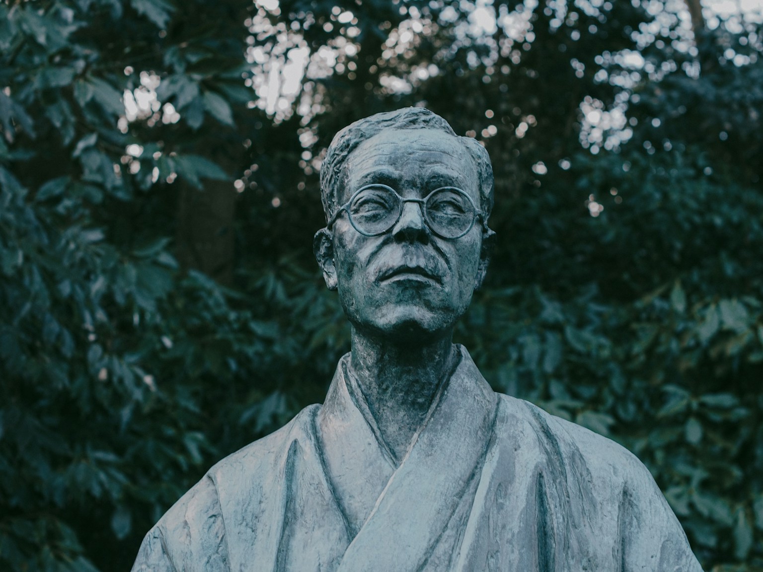 Patung perunggu seorang pria mengenakan kimono dan kacamata dengan latar belakang dedaunan hijau