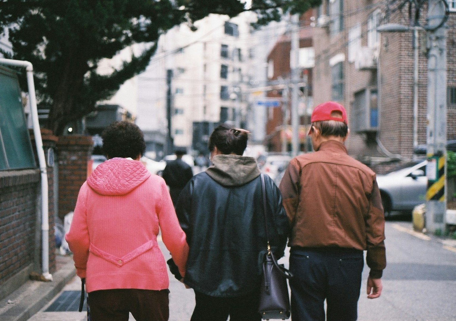 街を歩く三人の人々の後ろ姿 ピンクのジャケットを着た女性と黒いジャケットの男性と赤い帽子の男性