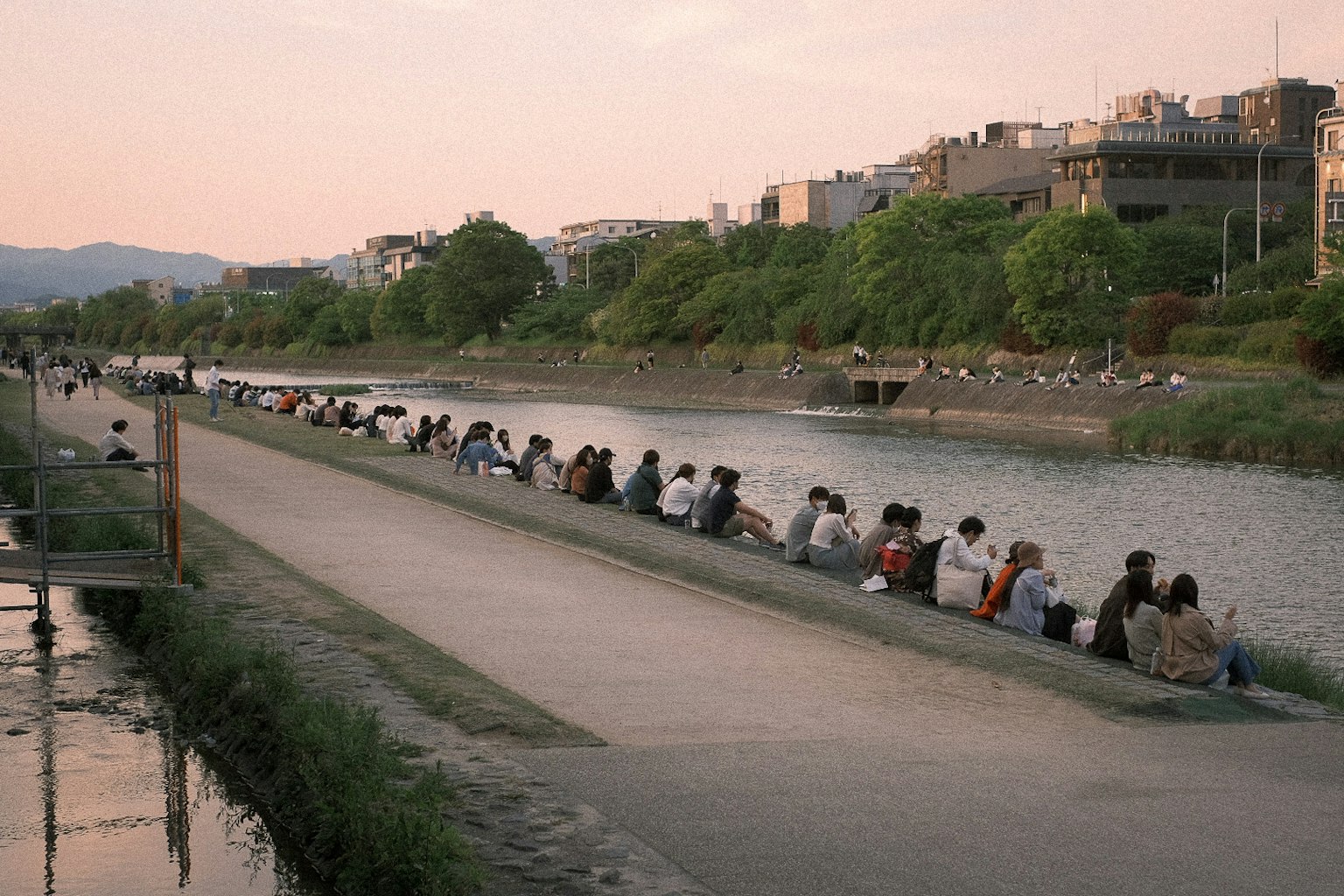 Fila di persone sedute lungo il fiume al tramonto con vegetazione e edifici sullo sfondo