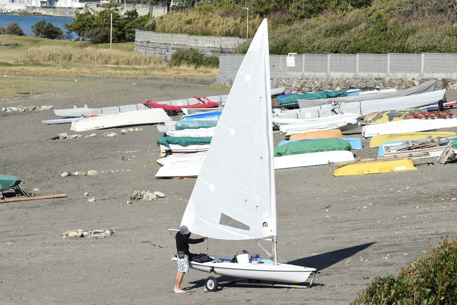 Mann bereitet ein kleines Segelboot am Strand mit einem weißen Segel bunte Boote im Hintergrund vor