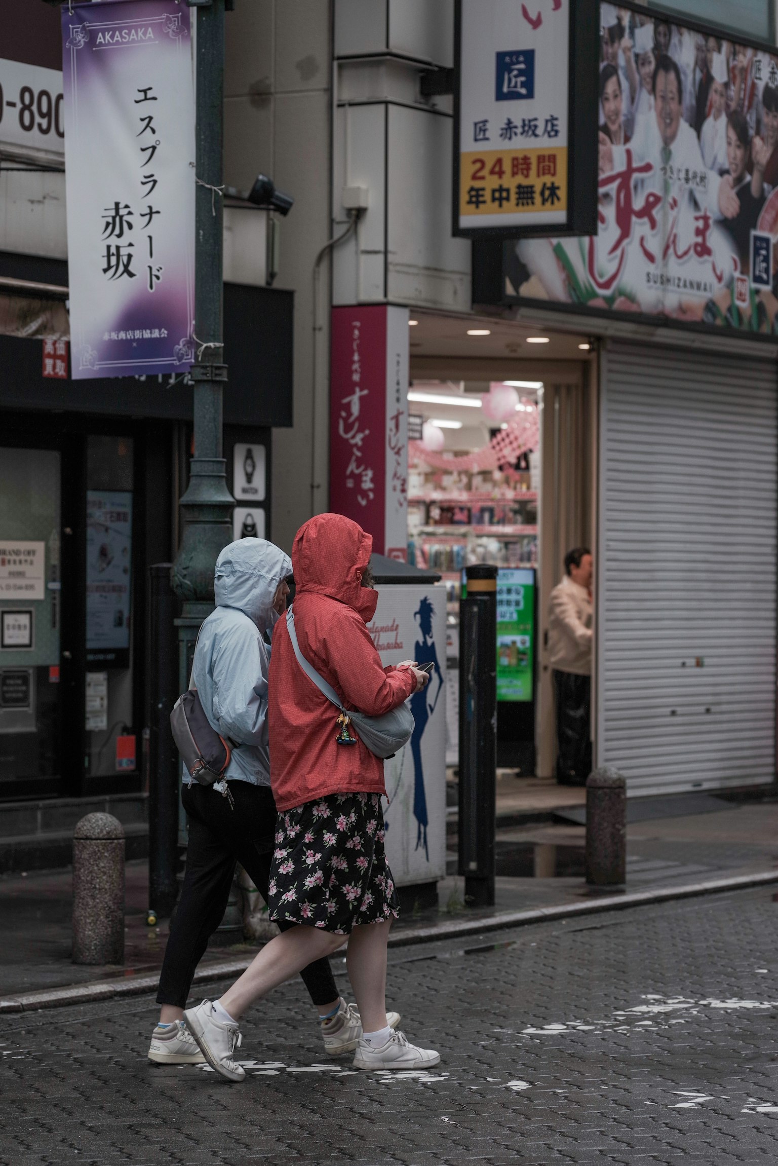 兩名女性在雨中行走 一人穿著藍色雨衣 另一人穿著紅色連帽外套 背景中可見商店招牌