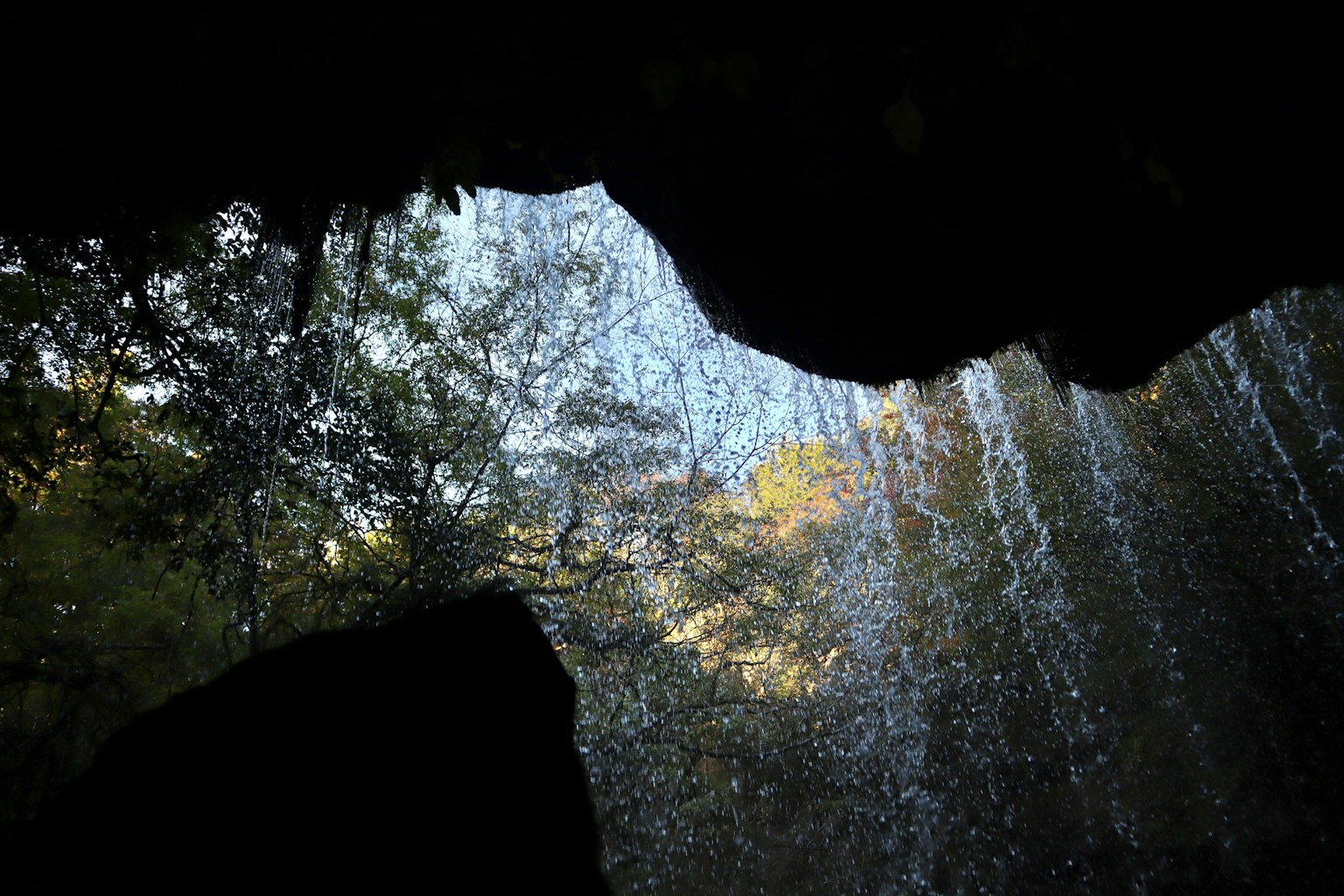 Pemandangan dari belakang air terjun dengan sinar matahari menerangi tetesan air