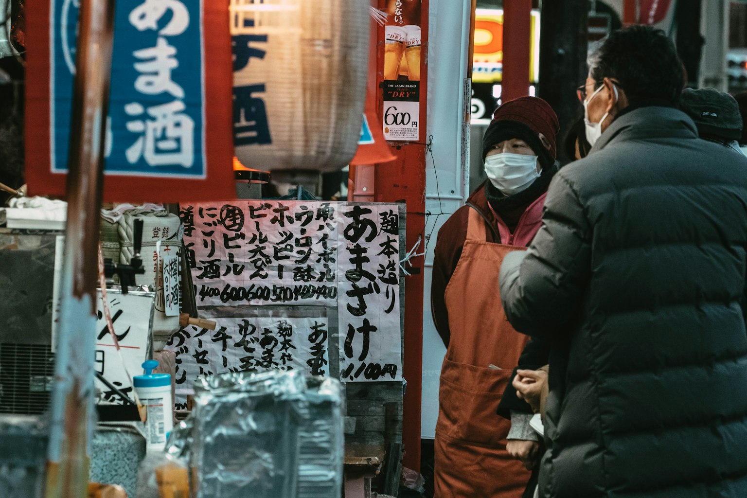 Escena nocturna en la calle con personas hablando frente a un puesto de comida y letreros japoneses