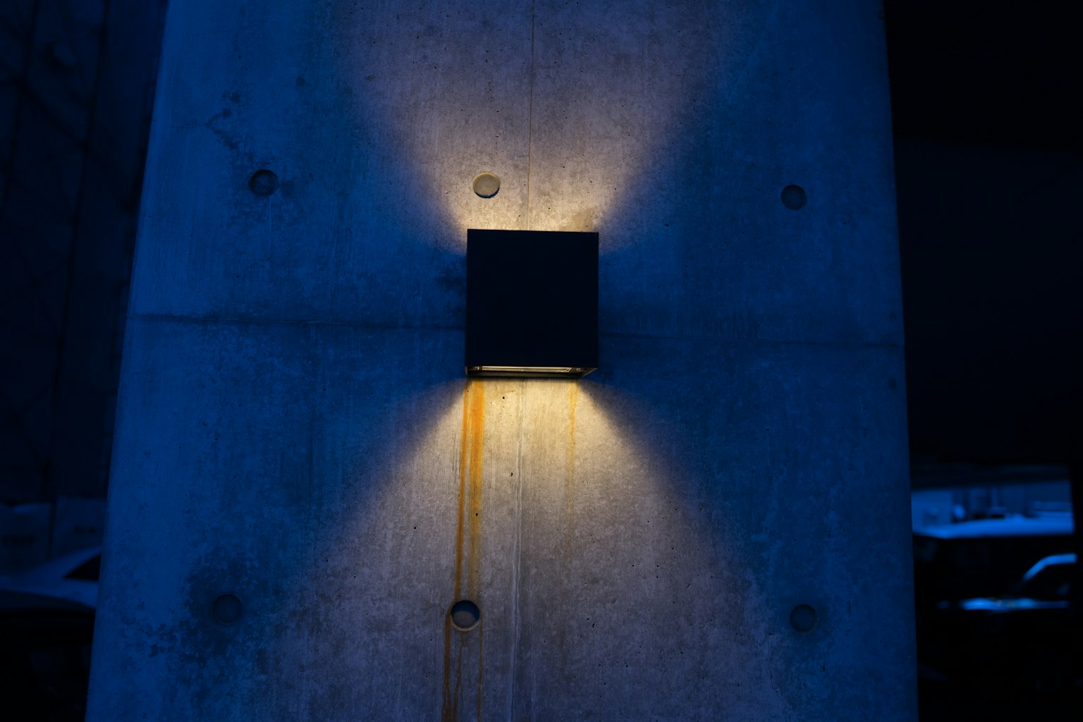 A square black light fixture illuminates a concrete wall in a dark setting