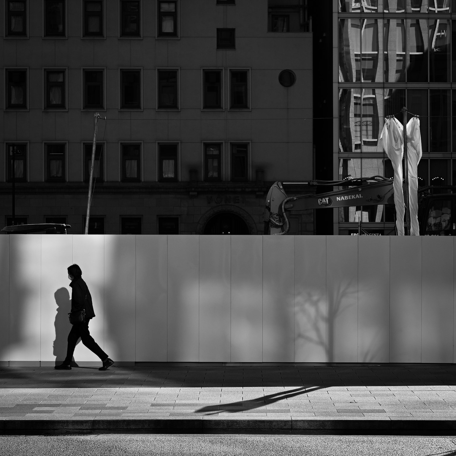Pemandangan kota hitam putih dengan seseorang yang berjalan, bayangan, dan bangunan serta patung mencolok di latar belakang
