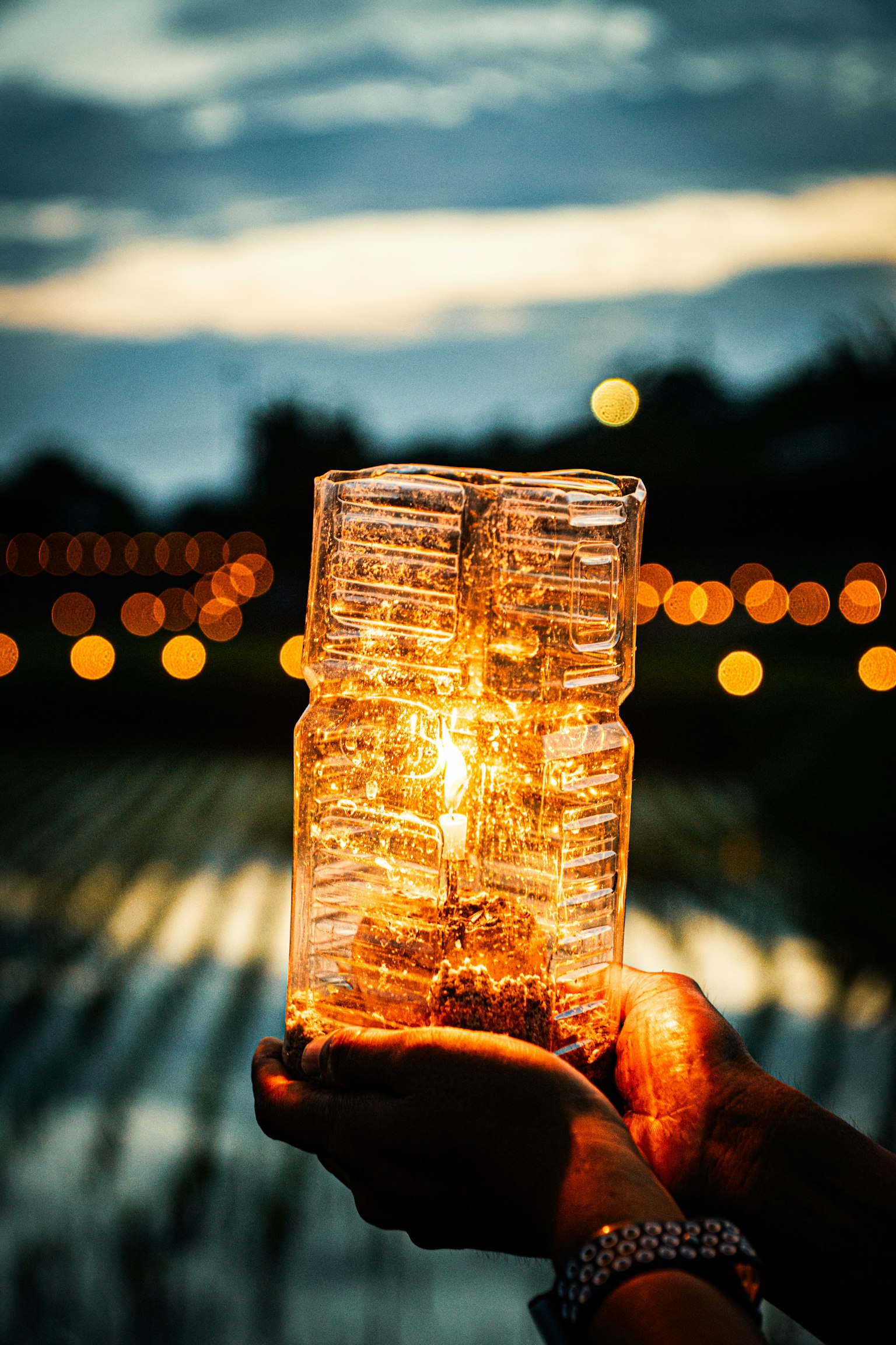 A glowing candle jar held in hands with a blurred background of lights