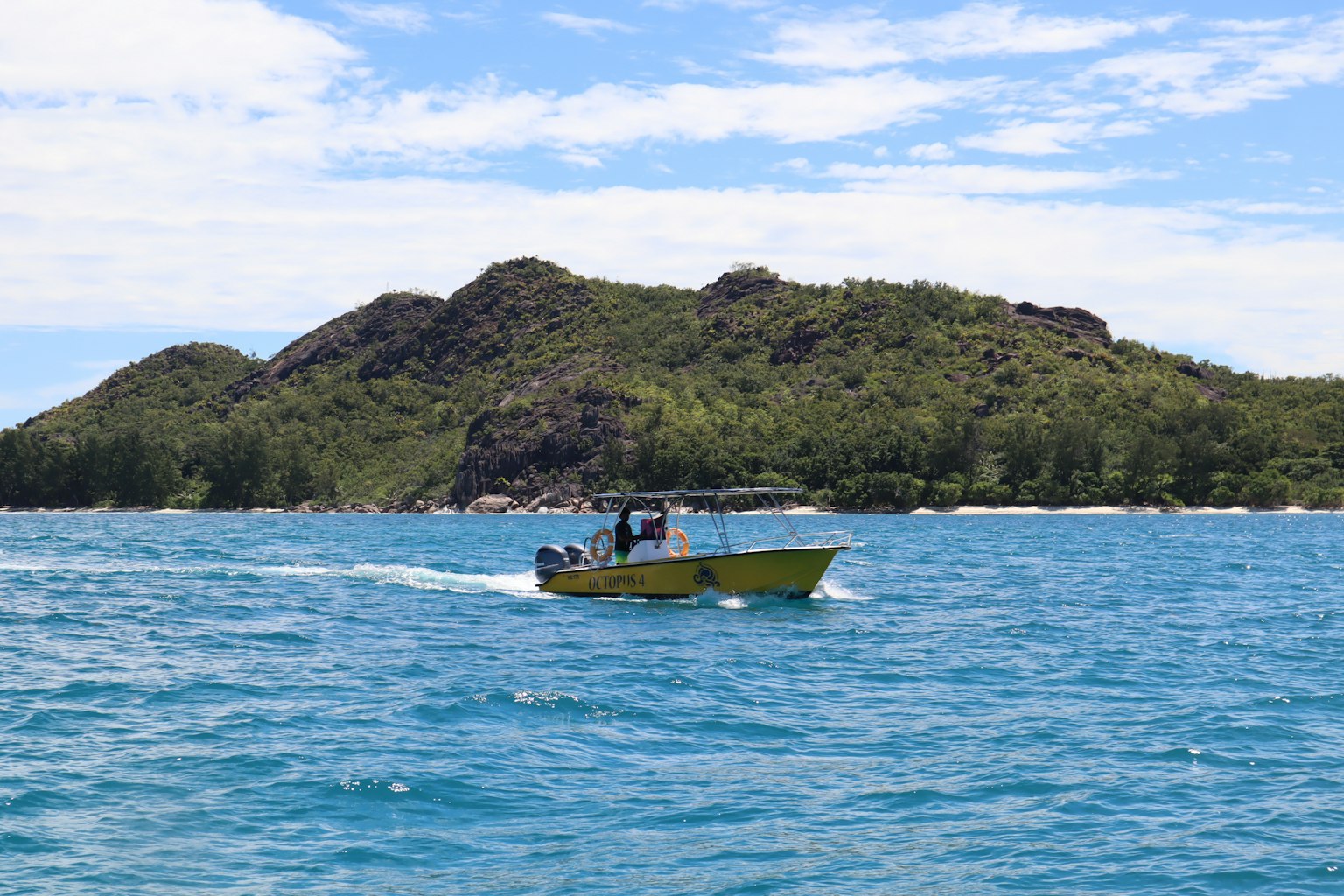 Perahu kuning berlayar di atas air biru dengan bukit hijau di latar belakang