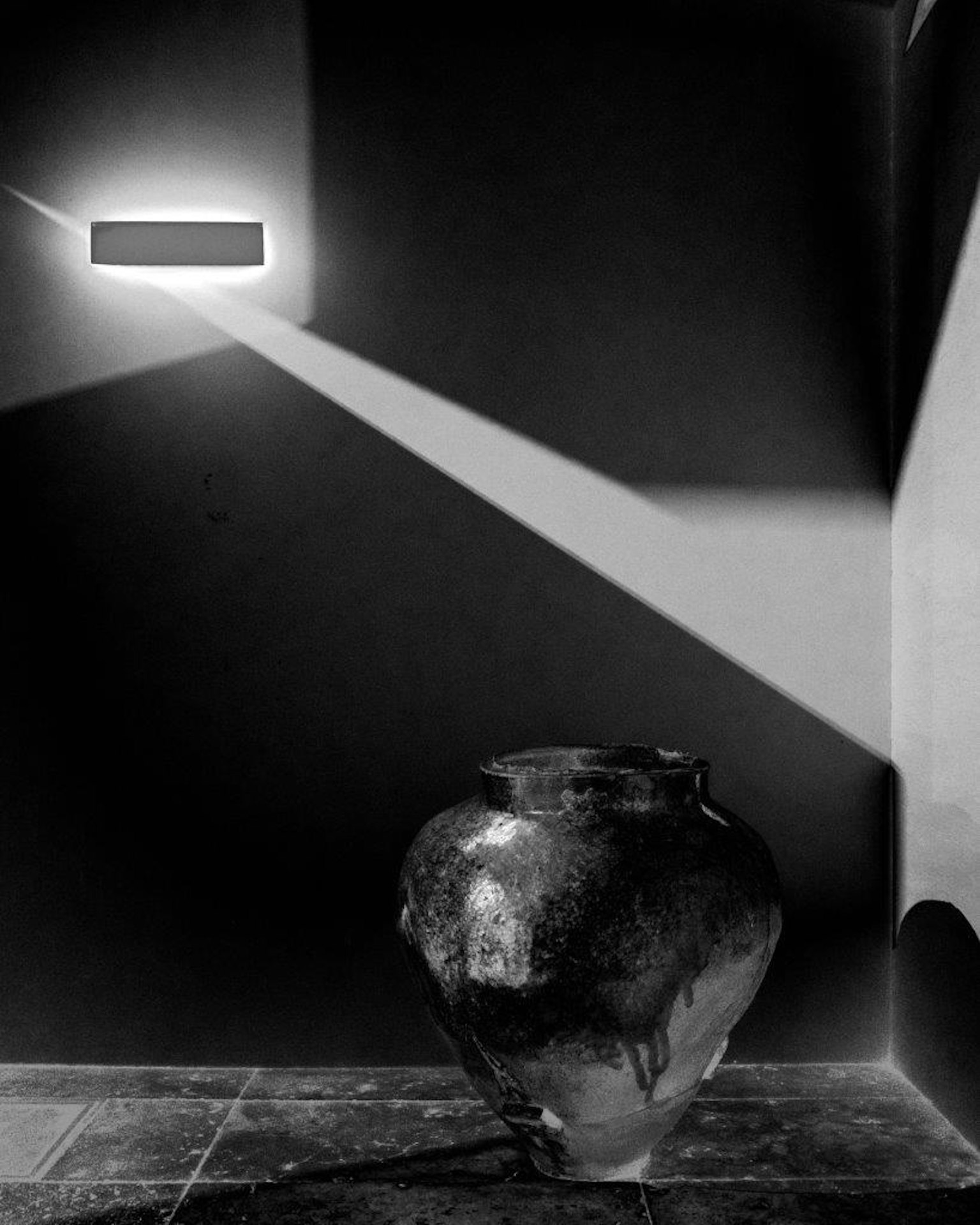 Photo en noir et blanc mettant en avant un pot en céramique dans le coin avec un faisceau de lumière diagonale provenant du mur