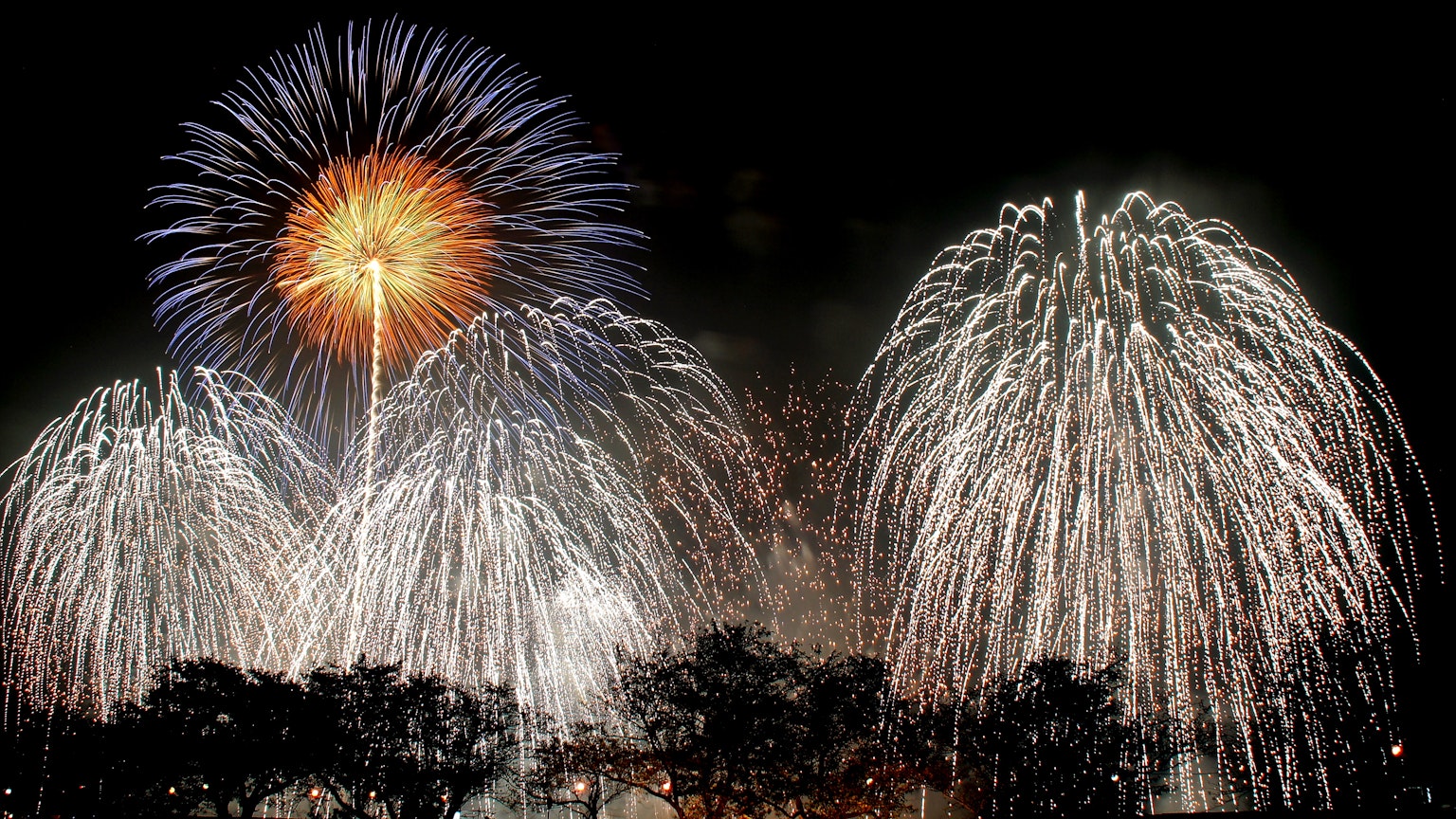 Vibrant fireworks display in the night sky featuring a large orange firework and cascading white fireworks