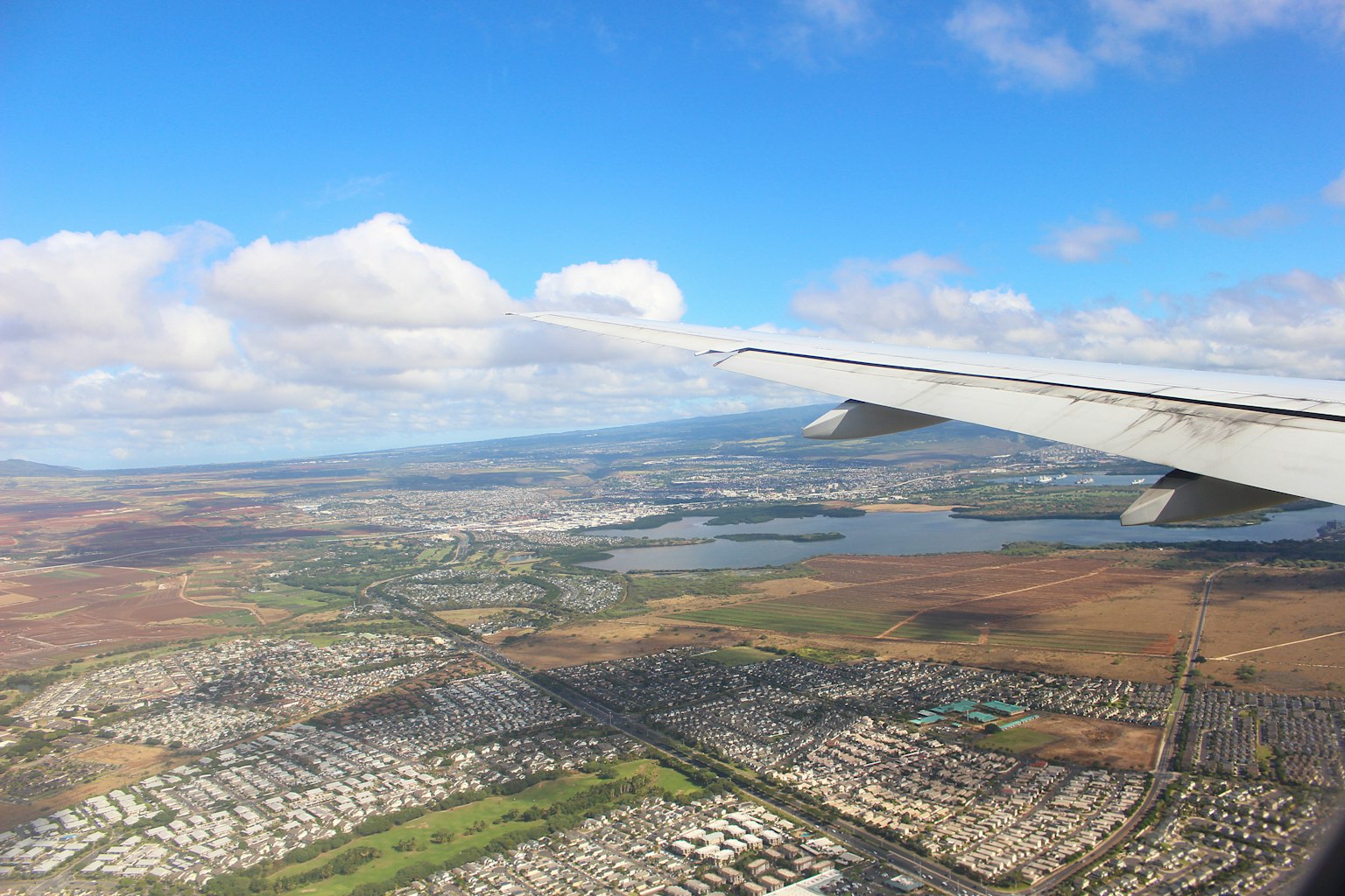 View from an airplane wing showing a landscape with blue sky and clouds, sprawling city and land